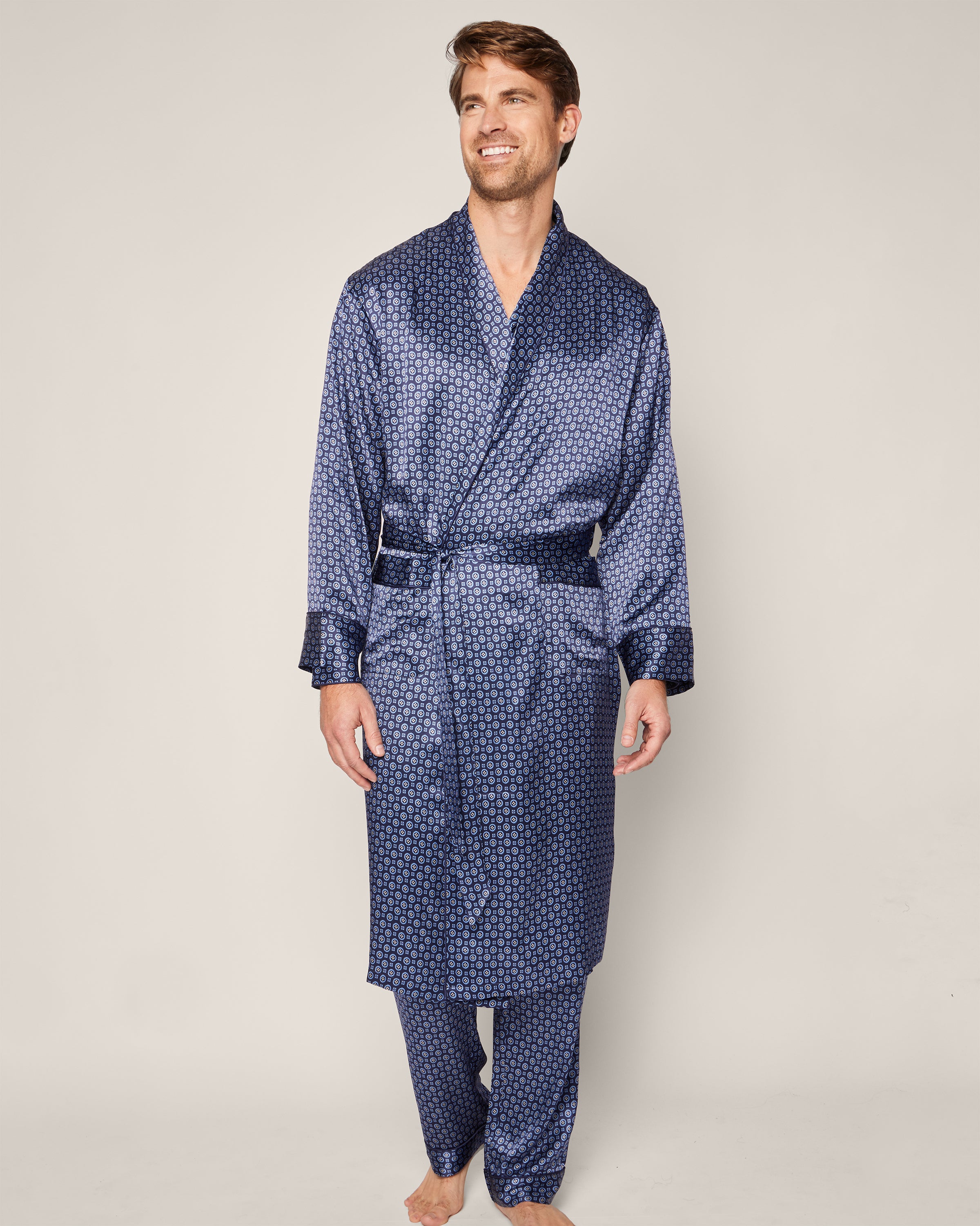 A person in the Petite Plume Mens Silk Robe in Midnight Foulard, a breathable blue silk robe with a geometric pattern and waist tie, is smiling barefoot against a light background, completing the elegant matching ensemble.