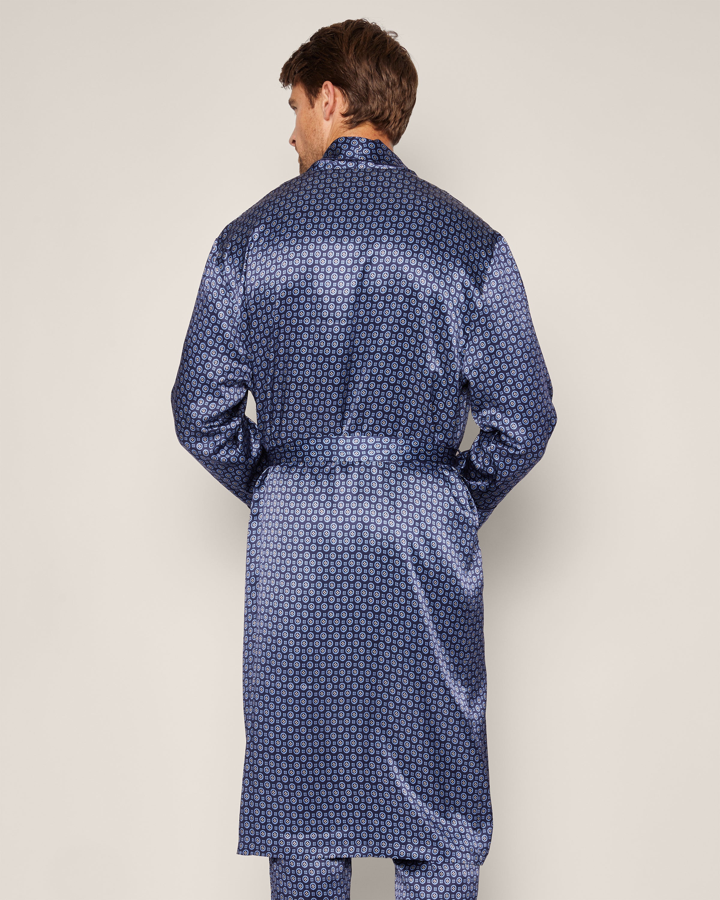 A man wearing Petite Plumes Mens Silk Robe in Midnight Foulard, featuring a subtle circular pattern and matching belt, stands with his back to the camera against a light background. The shiny, dark blue robe exemplifies breathable luxury sleepwear.