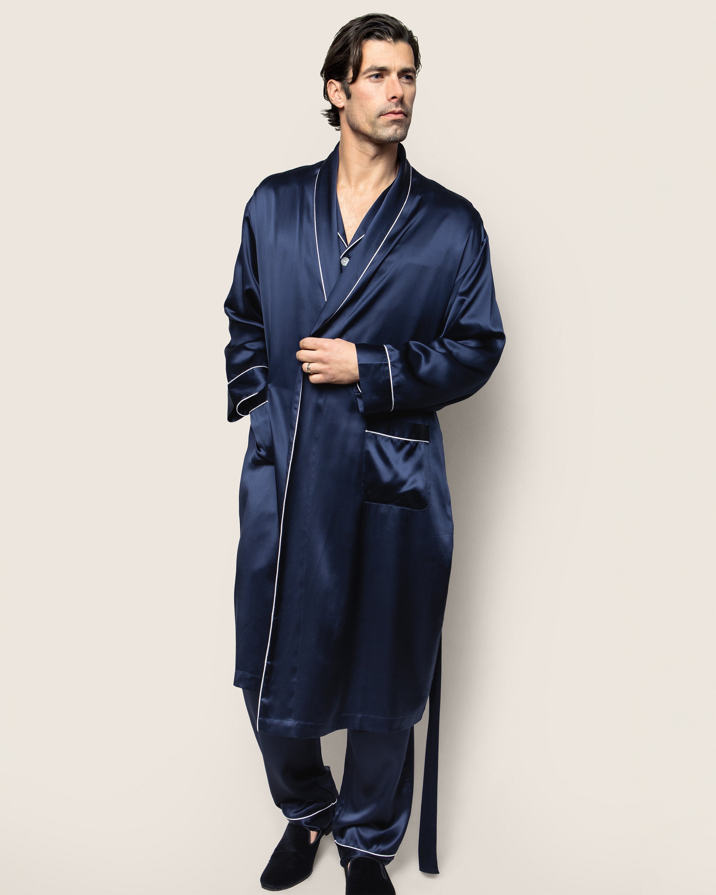A man with dark hair stands against a neutral background, wearing Petite Plumes Mens Silk Long Robe in Navy. Made from luxurious mulberry silk, the effortlessly chic robe is part of a sleepwear set paired with matching pants, while he gazes to the side with one hand in his pocket.