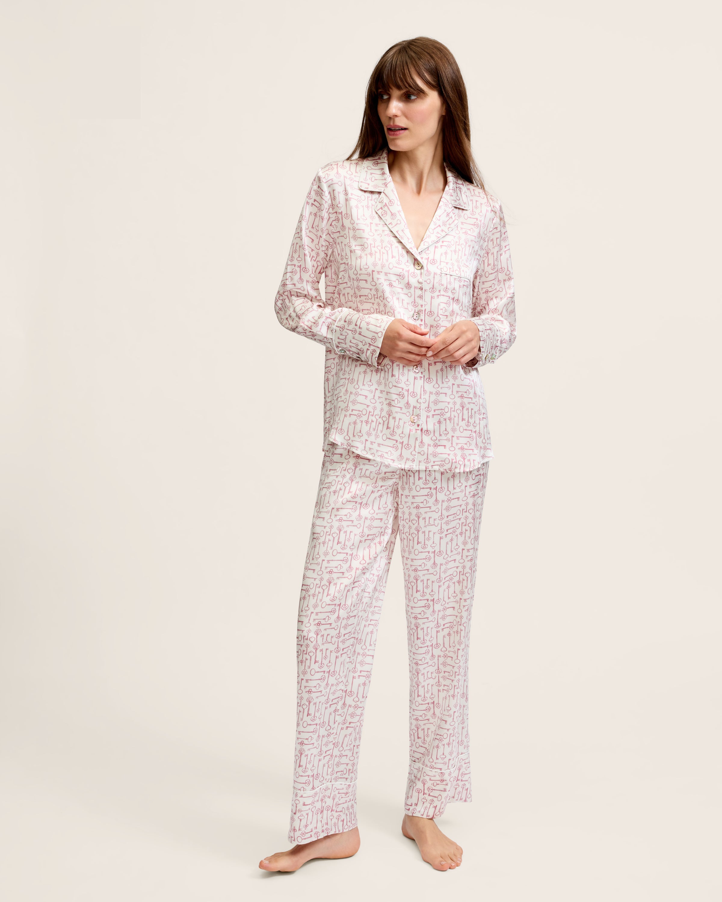 The Women's Silk Pajama Set in Key to My Heart features long-sleeve, button-up pajamas and matching pants in white silk with a pink wrench pattern, displayed flat on a white background.