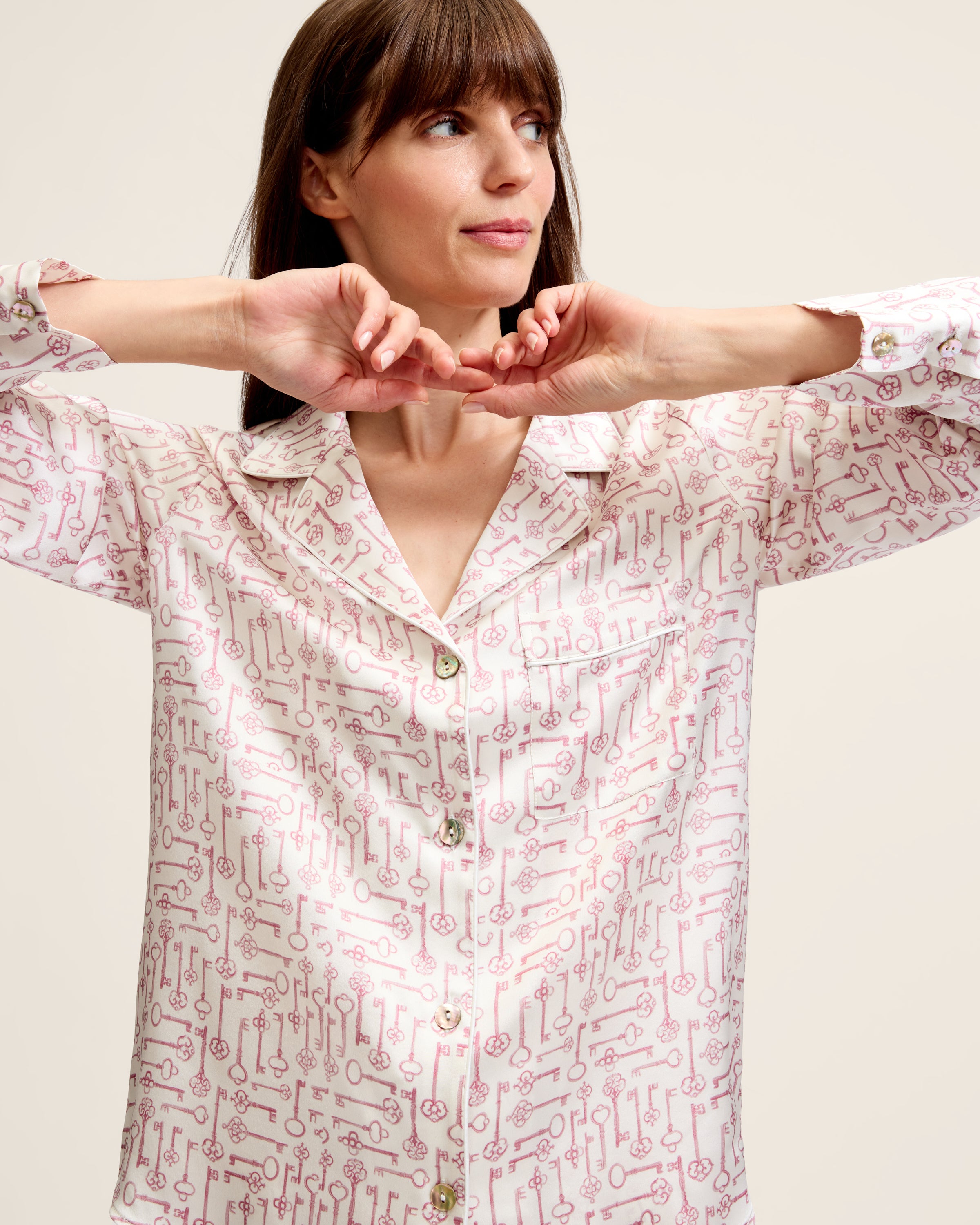 The Women's Silk Pajama Set in Key to My Heart features long-sleeve, button-up pajamas and matching pants in white silk with a pink wrench pattern, displayed flat on a white background.