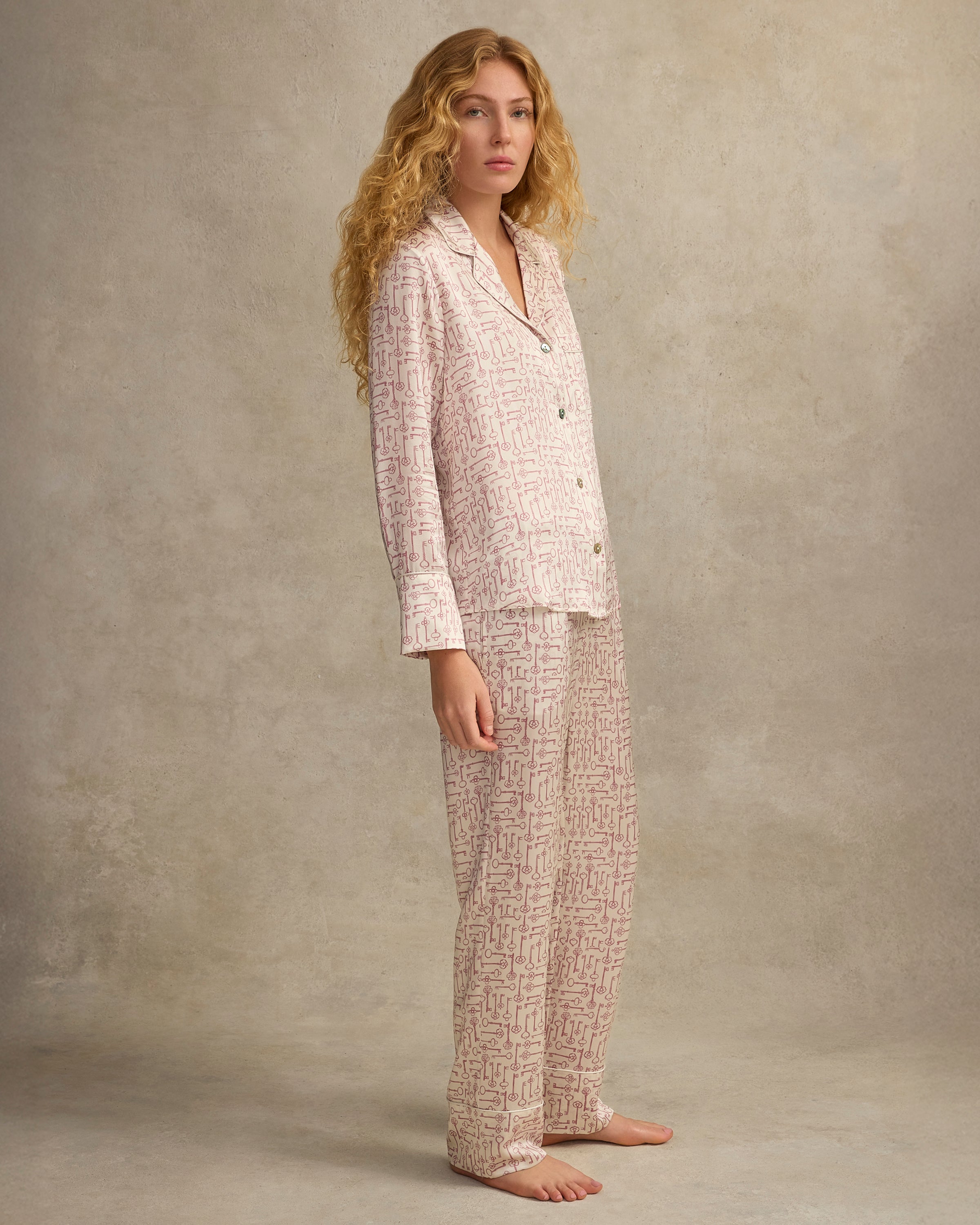 The Women's Silk Pajama Set in Key to My Heart features long-sleeve, button-up pajamas and matching pants in white silk with a pink wrench pattern, displayed flat on a white background.