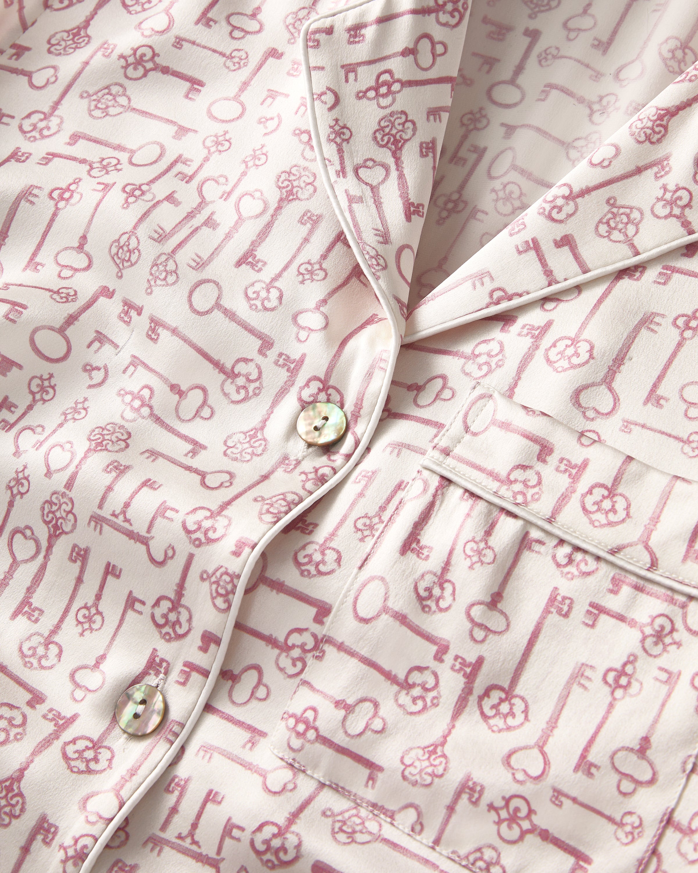 The Women's Silk Pajama Set in Key to My Heart features long-sleeve, button-up pajamas and matching pants in white silk with a pink wrench pattern, displayed flat on a white background.