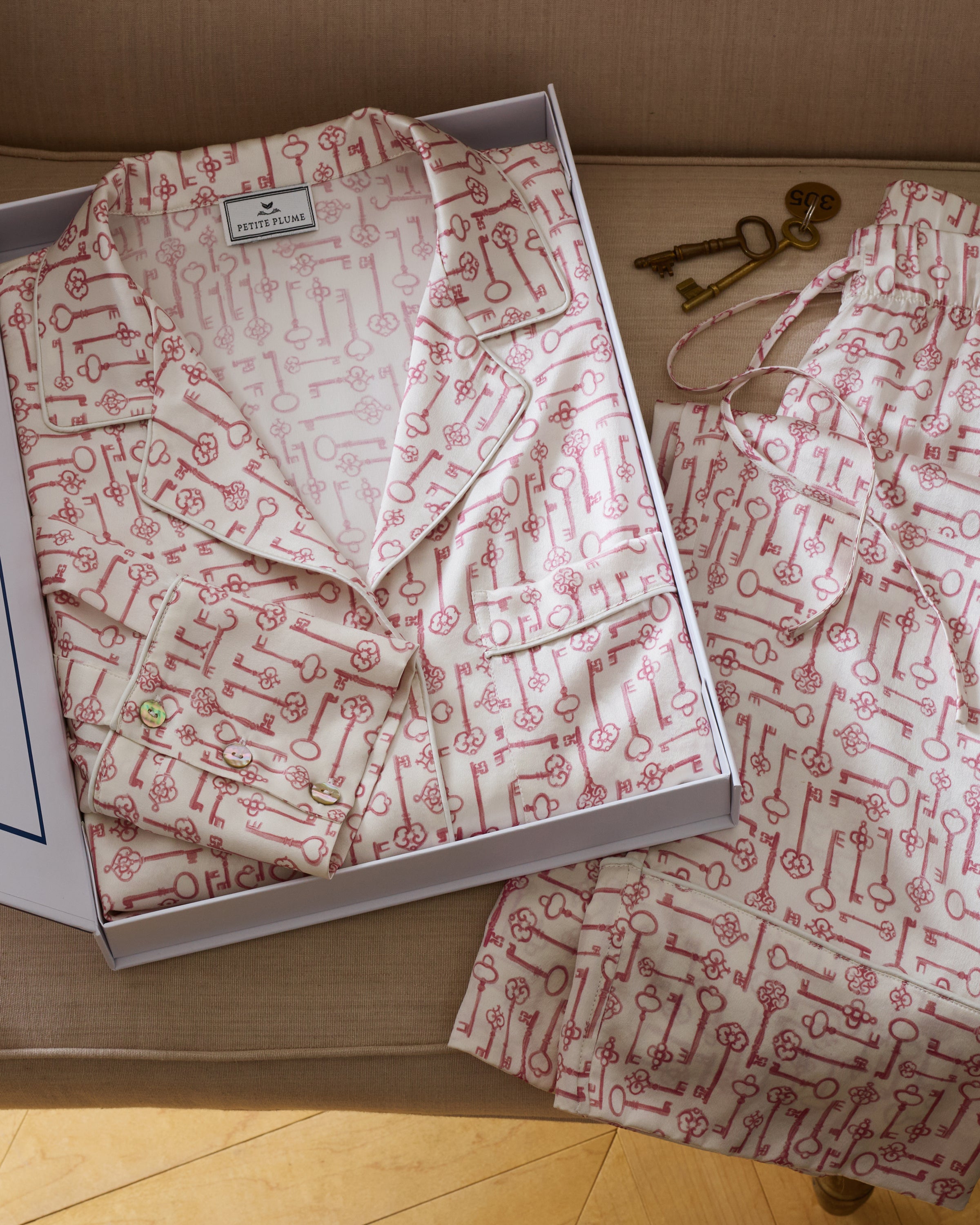 The Women's Silk Pajama Set in Key to My Heart features long-sleeve, button-up pajamas and matching pants in white silk with a pink wrench pattern, displayed flat on a white background.