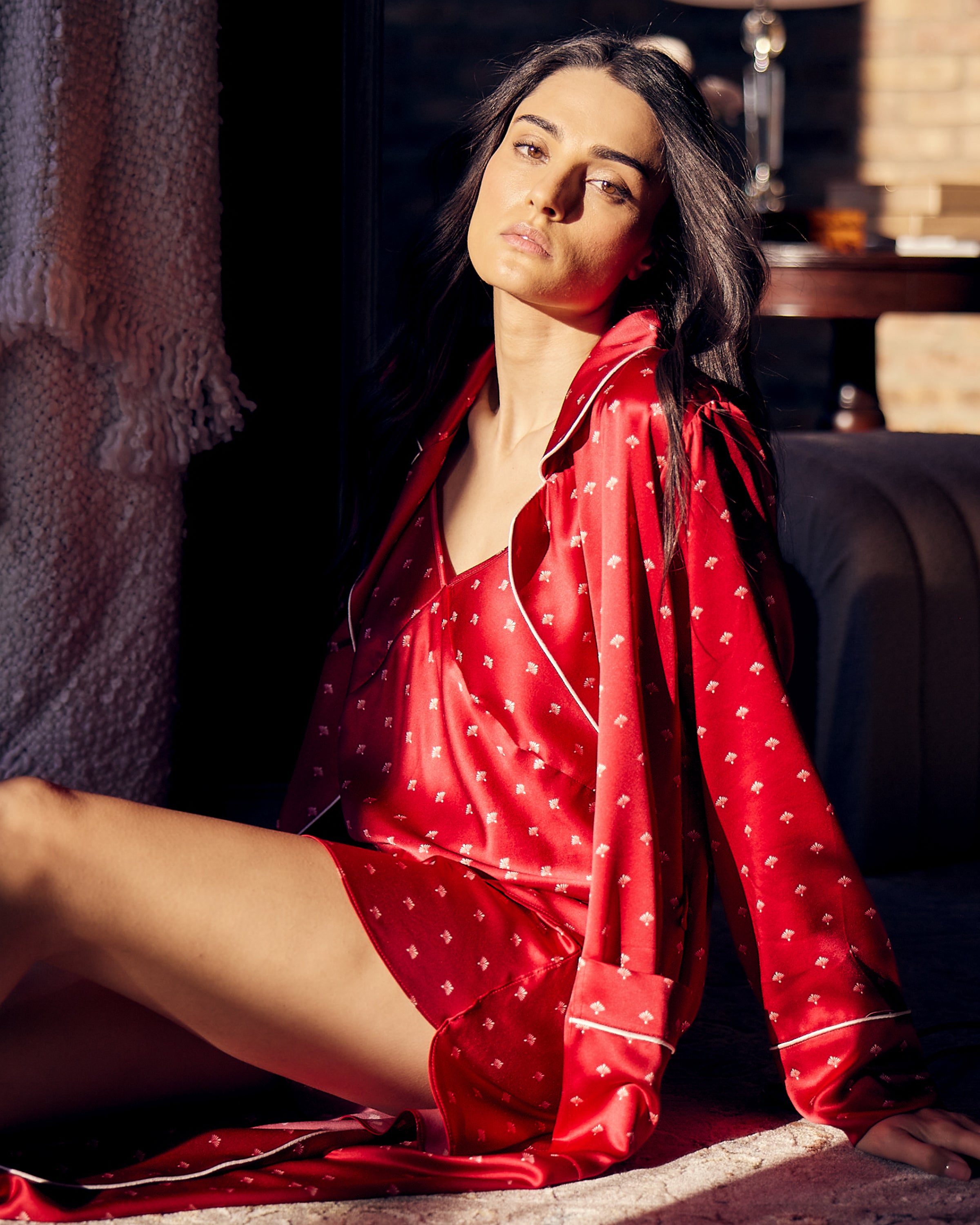 A person with long dark hair sits on the floor wearing Petite Plumes Womens Silk Cami Short Set in Crimson Art Nouveau. Sunlight streams in, casting shadows. They appear relaxed, leaning against a couch with a cozy blanket and Art Nouveau print bookshelves behind them.
