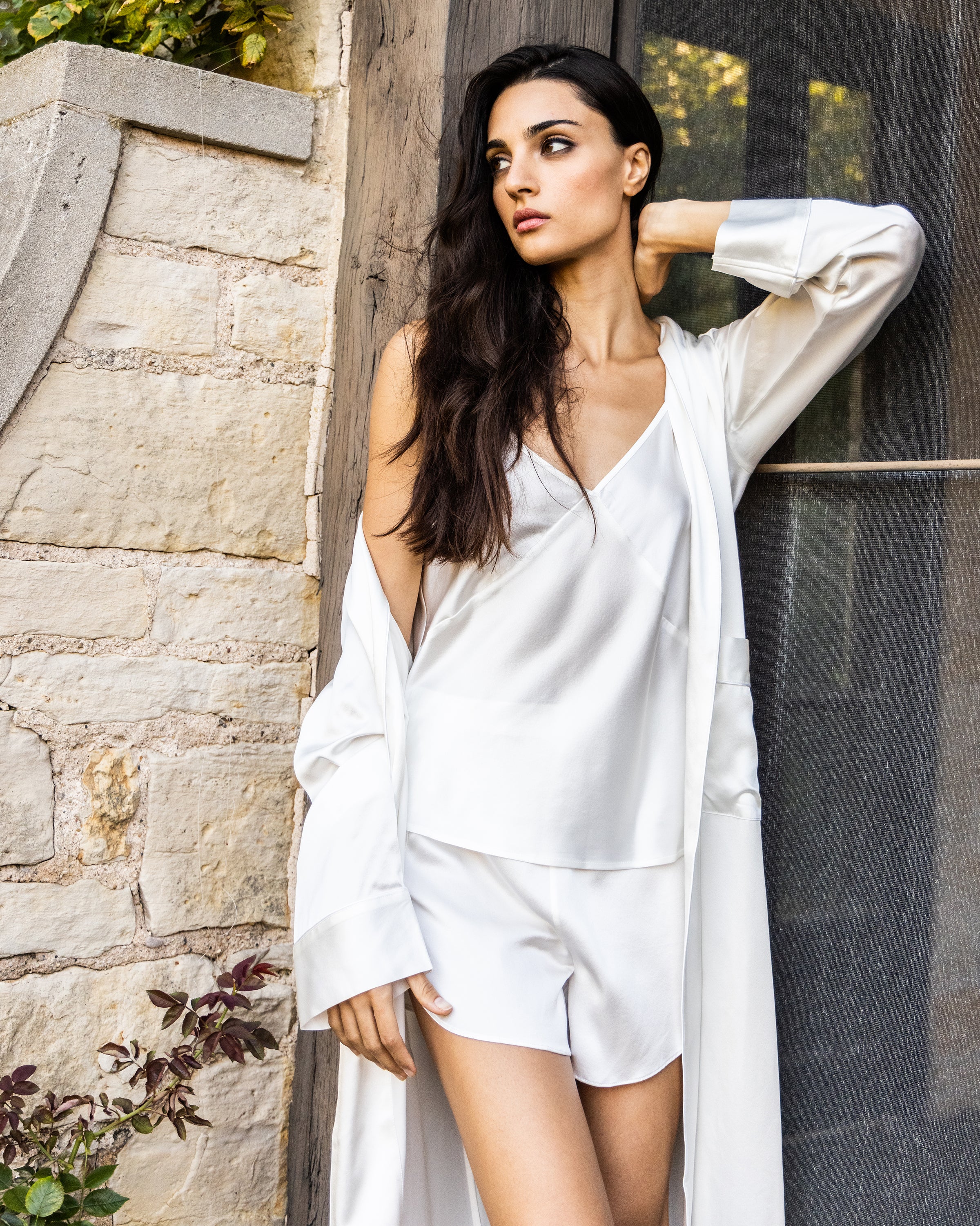 Wearing Petite Plumes Womens Silk Cami Short Set in White, a woman leans against a wooden and stone wall. Her long hair rests over one shoulder as she gazes thoughtfully to the side, with a relaxed setting and partial foliage visible at the top corner.