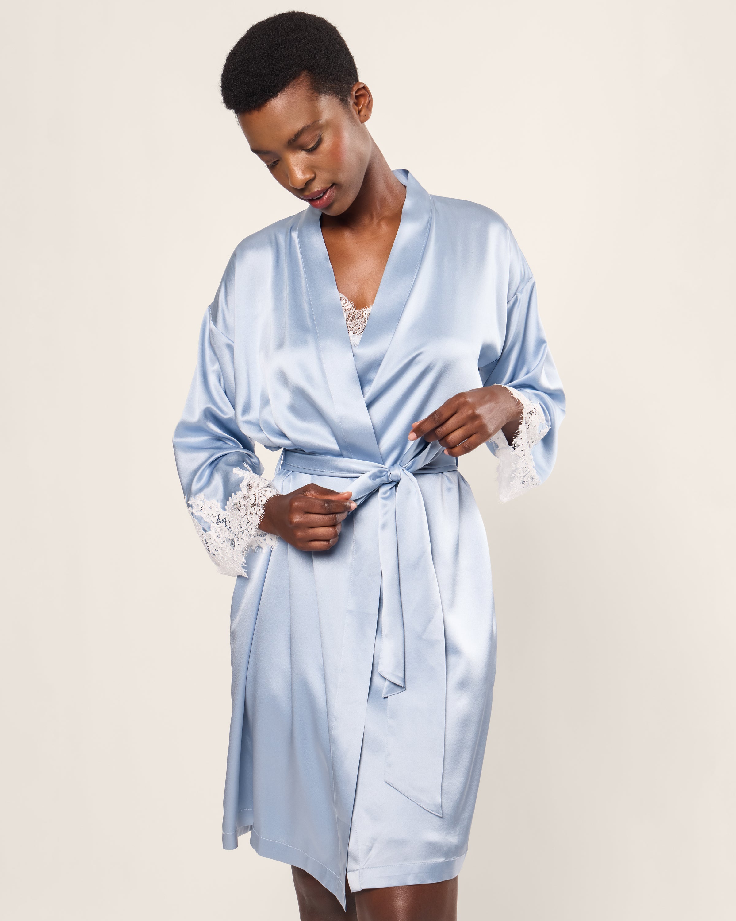 A person in a Petite Plume Womens Silk Lace Robe in Periwinkle stands against a plain background, adjusting the belt to reveal lace beneath. Crafted from mulberry silk, this elegant and hypoallergenic sleepwear offers luxury and comfort.