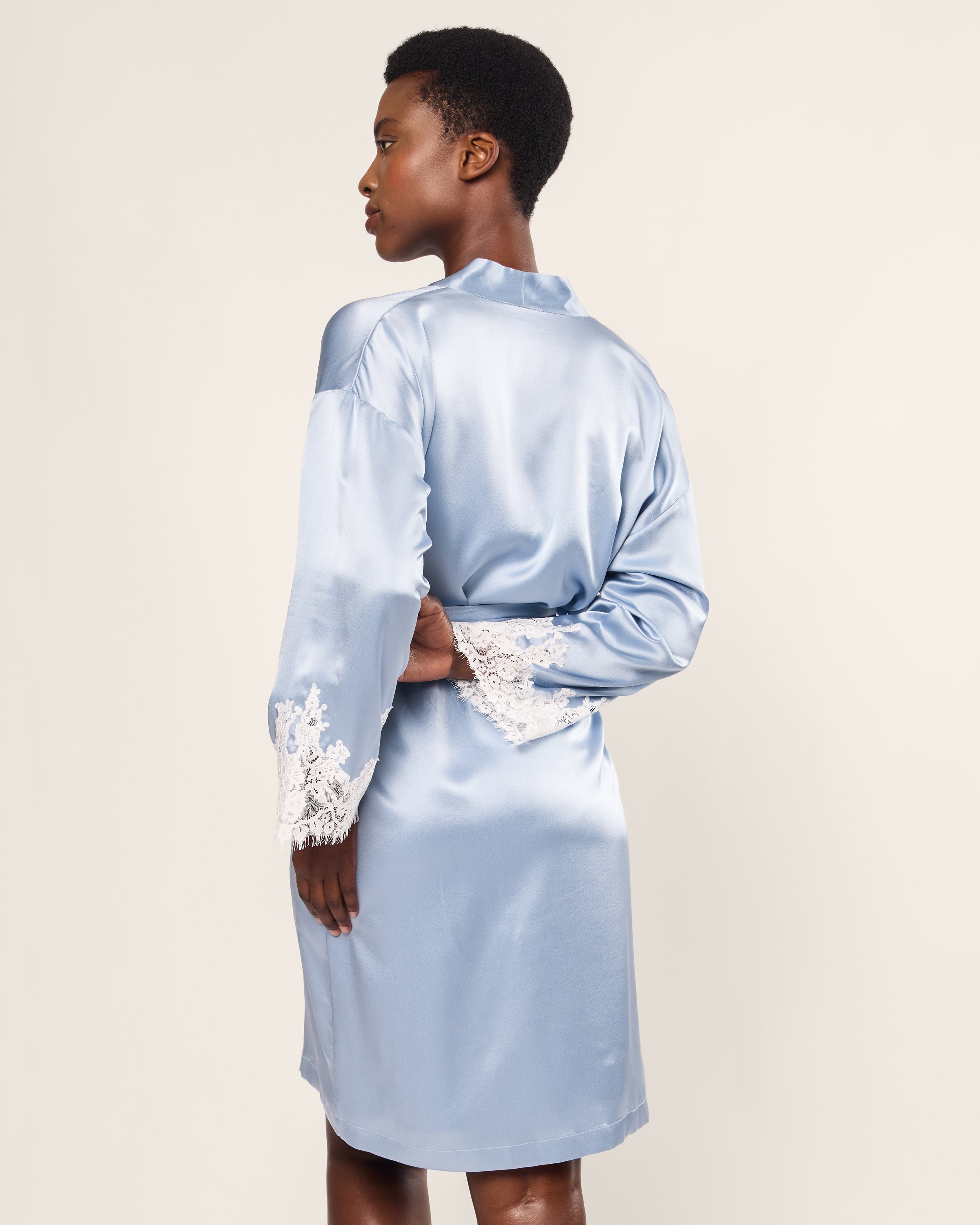 Standing with their back to the camera, a person wears Petite Plumes Womens Silk Lace Robe in Periwinkle, notable for its Mulberry Silk fabric and white lace cuffs. Their short hair and gentle pose highlight the robes elegance against a light backdrop.