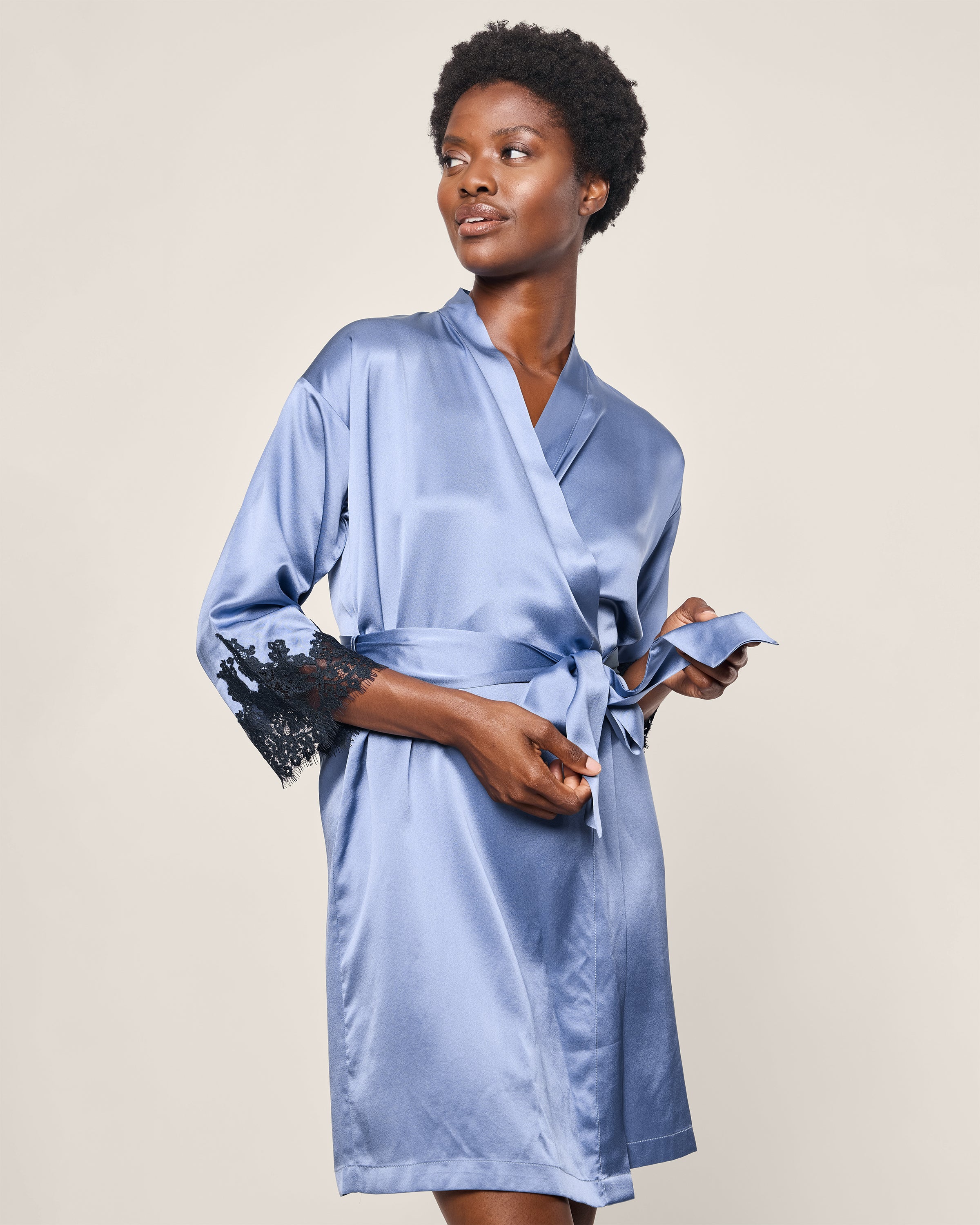 Dressed in Petite Plumes Womens Silk Lace Robe in Mystique Blue, a person poses against a plain background. The mulberry silk robe features black lace-trimmed sleeves and is cinched at the waist with a matching satin belt.