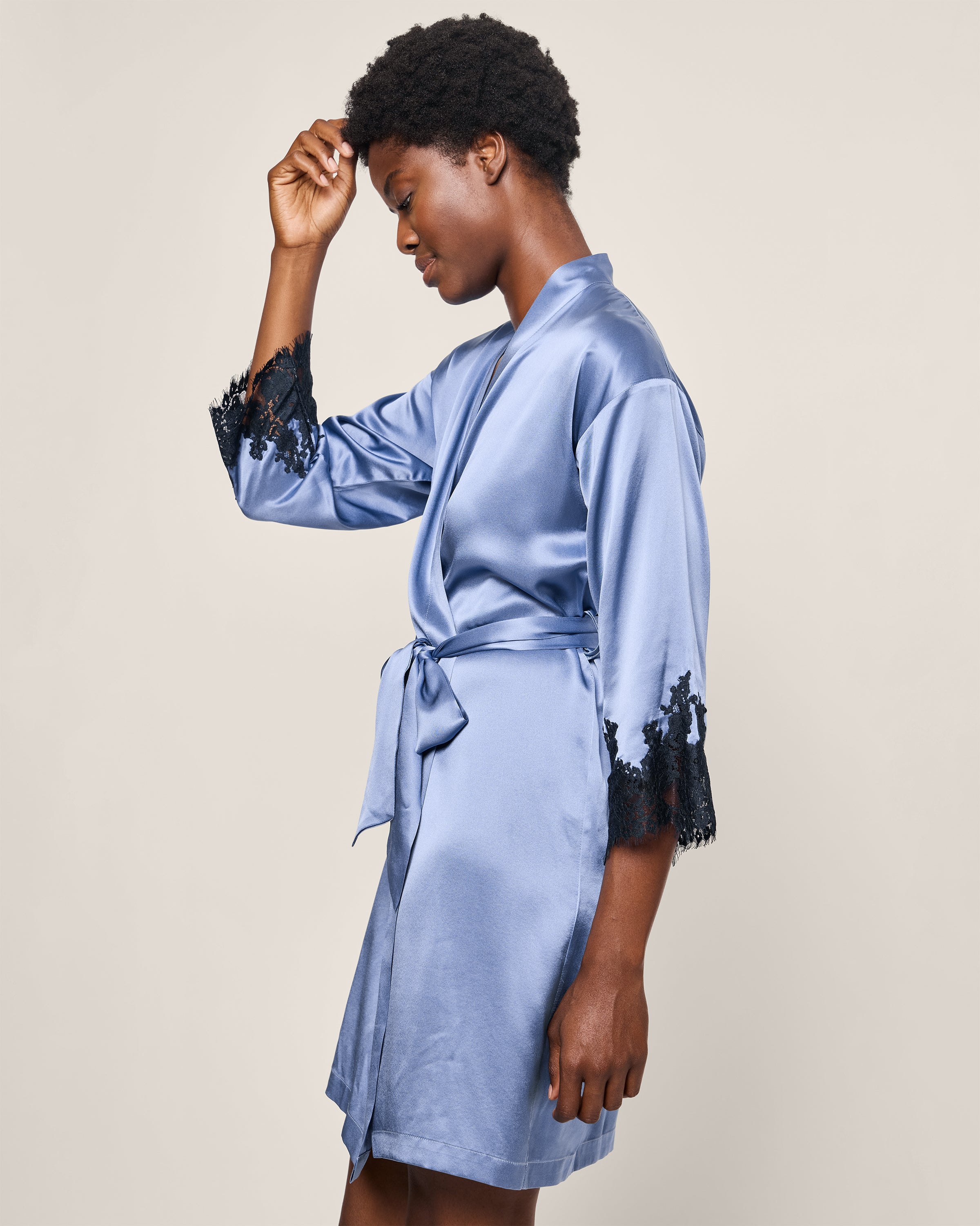 Against a plain backdrop, a person in profile wears the Womens Silk Lace Robe in Mystique Blue by Petite Plume. They gracefully place their hand near their forehead, highlighting the luxurious tie and hypoallergenic fabric of this mulberry silk sleepwear with black lace trim.
