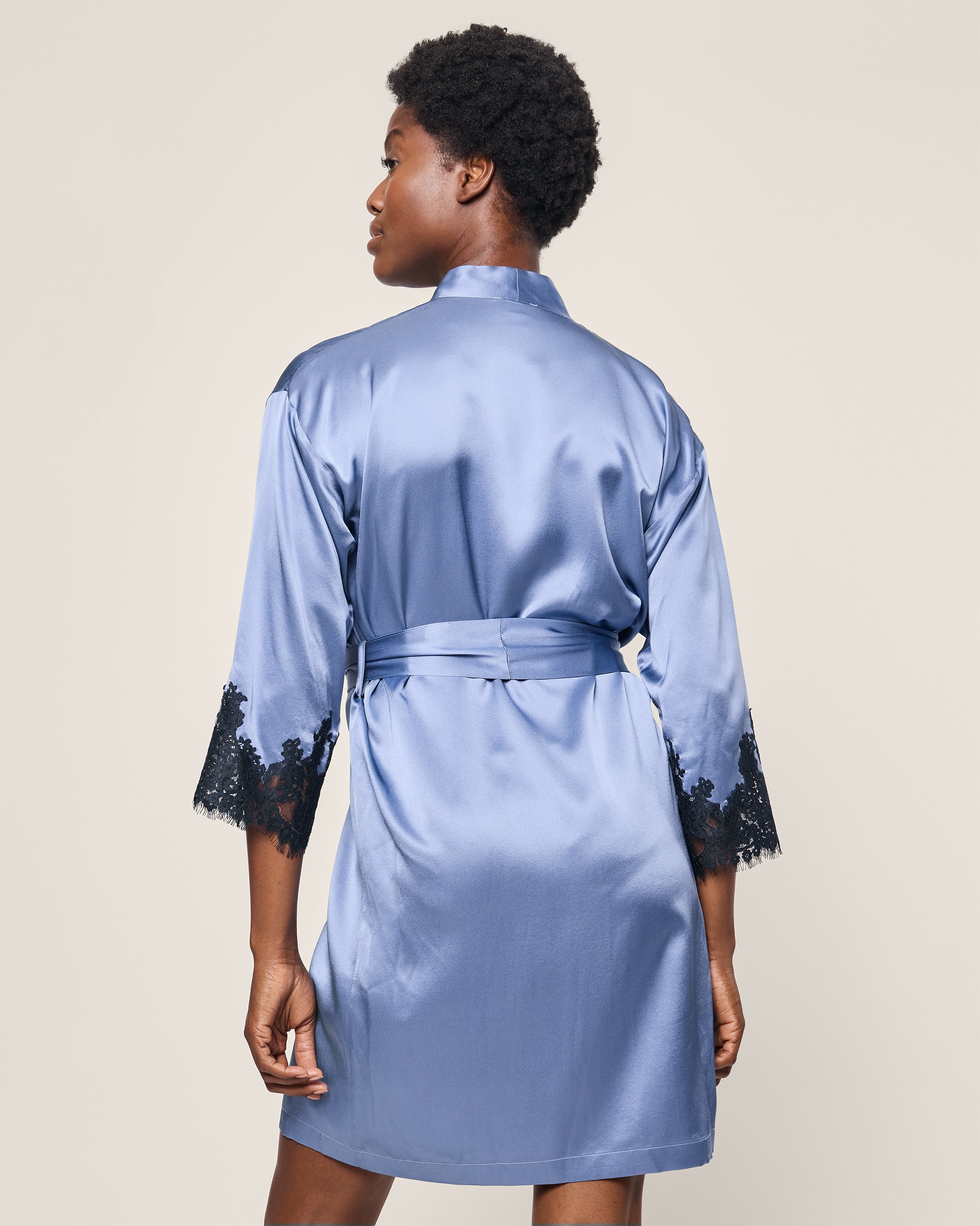 Facing away from the camera, a person wears Petite Plumes Womens Silk Lace Robe in Mystique Blue, showcasing luxurious hypoallergenic mulberry silk with black lace sleeve trim. The robe is cinched at the waist against a plain light-colored background.
