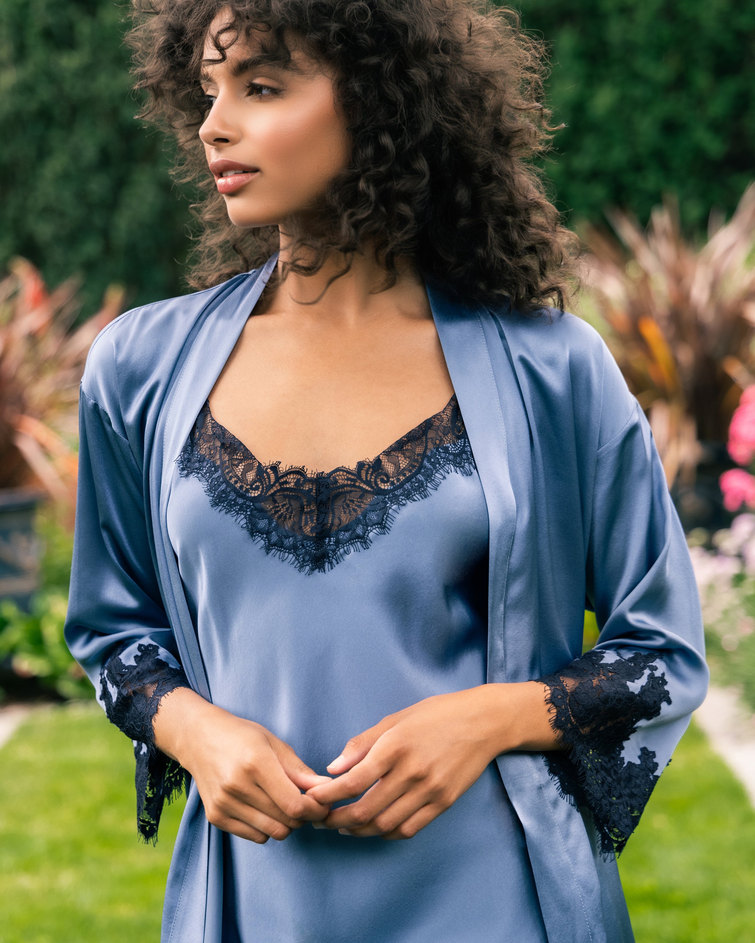 A woman with curly hair wears a Petite Plume Womens Silk Lace Robe in Mystique Blue, standing in a garden. She gazes to the side, surrounded by colorful plants and greenery.