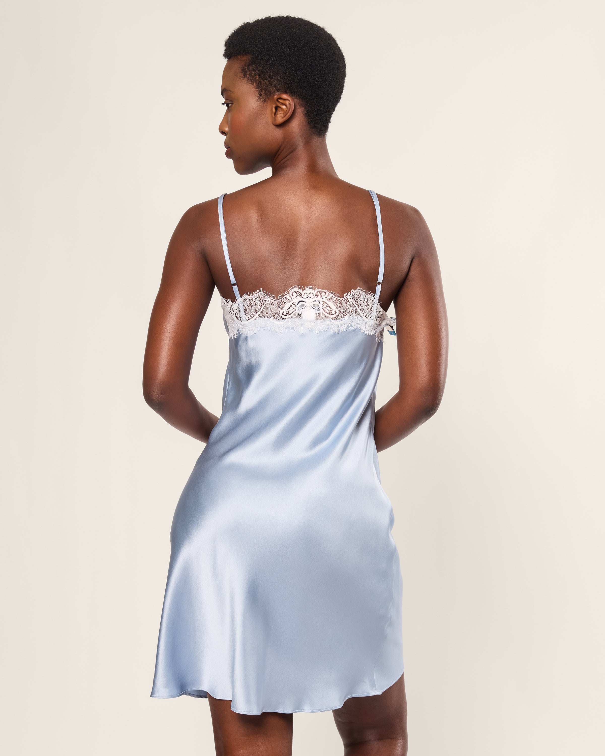 A person in Petite Plumes Womens Silk Lace Slip Dress in Periwinkle with delicate detailing and thin straps stands with their back to the camera. The luxurious slip, ending above the knees, is set against a plain, light-colored background.