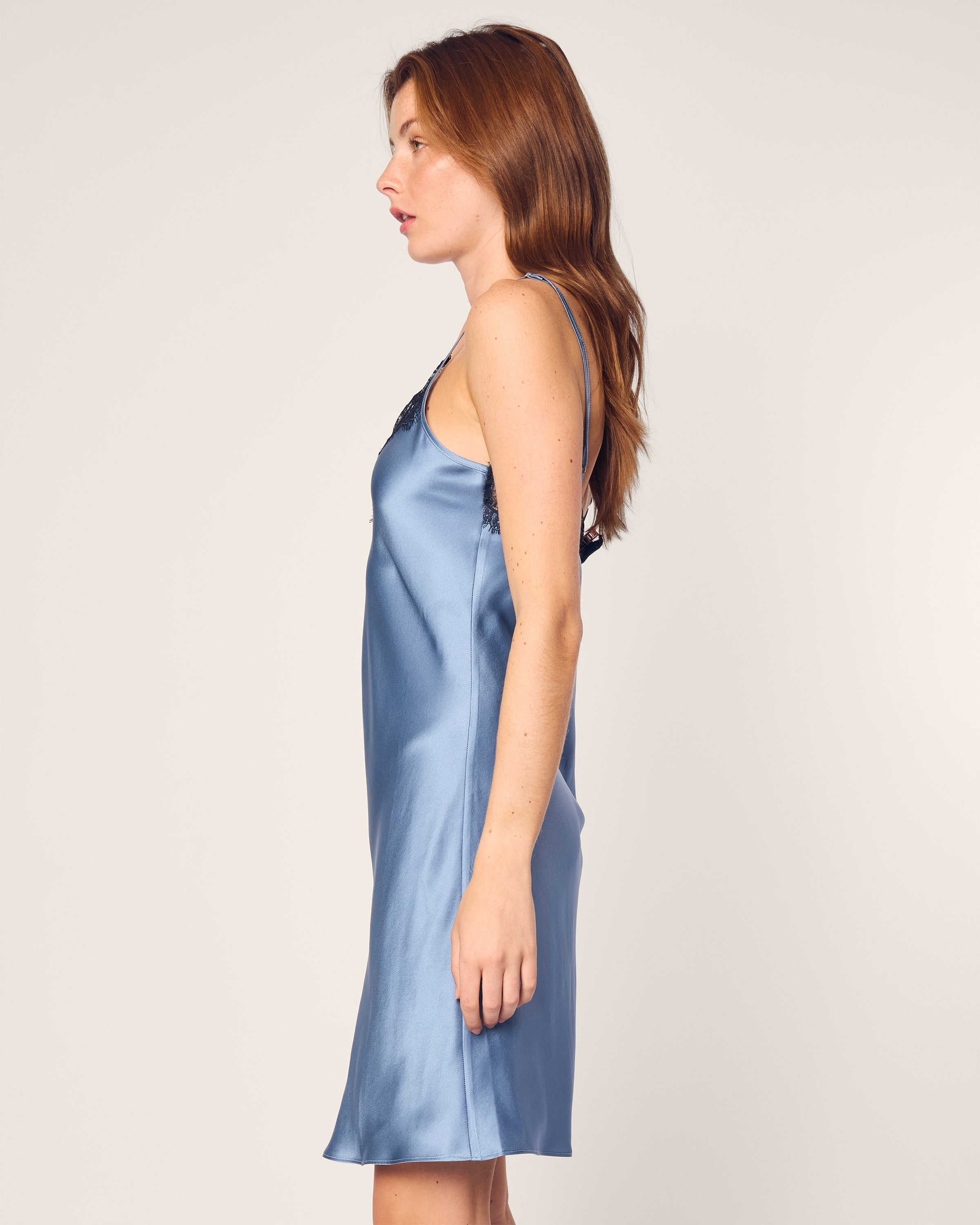 A person with long auburn hair stands in profile, wearing the Womens Silk Lace Slip Dress in Mystique Blue by Petite Plume, featuring black lace trim. The background is plain and off-white.
