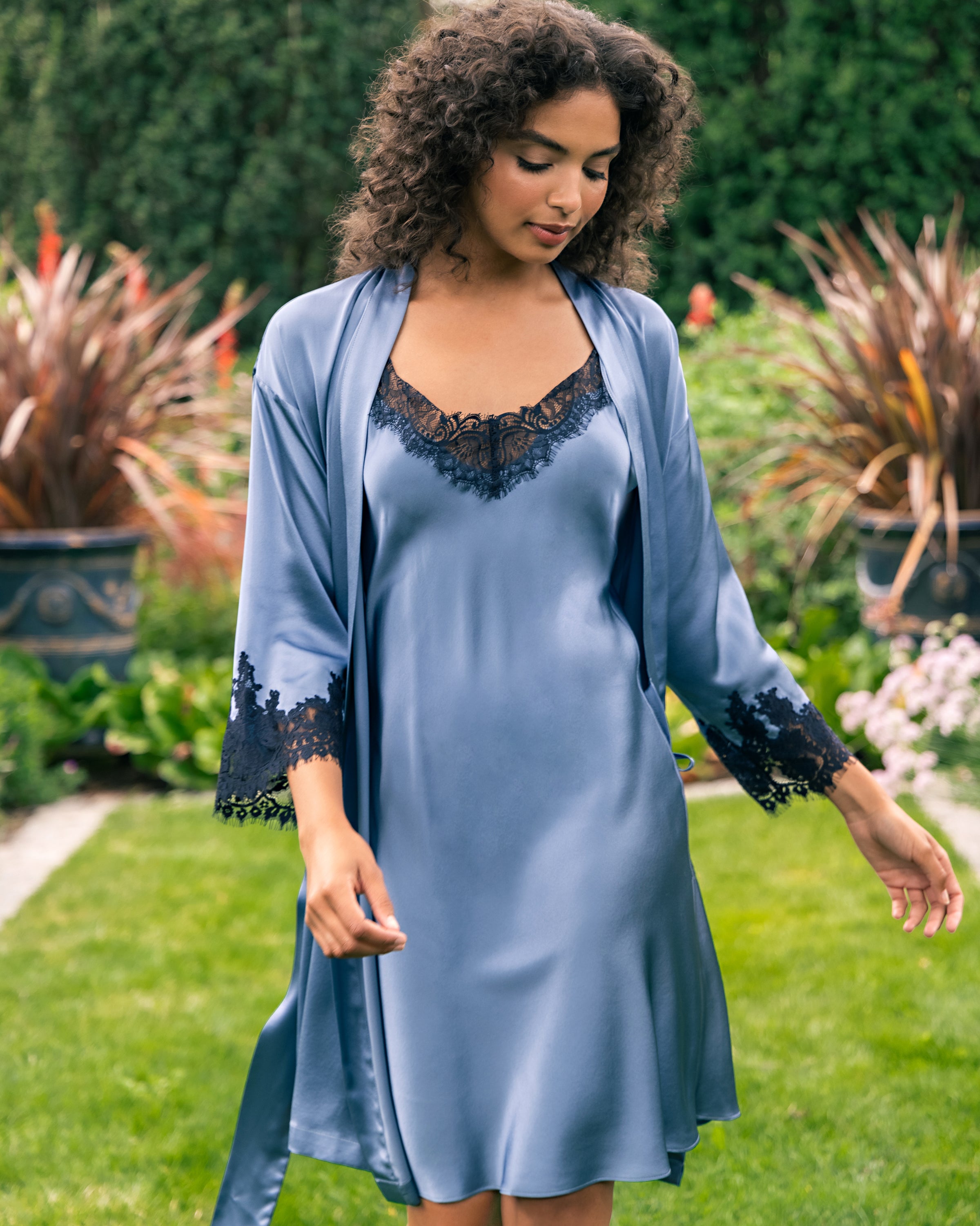Amid lush greenery, a person with curly hair embodies minimalist sensuality in Petite Plumes Womens Silk Lace Slip Dress in Mystique Blue. They look down, clutching the dresss belt.