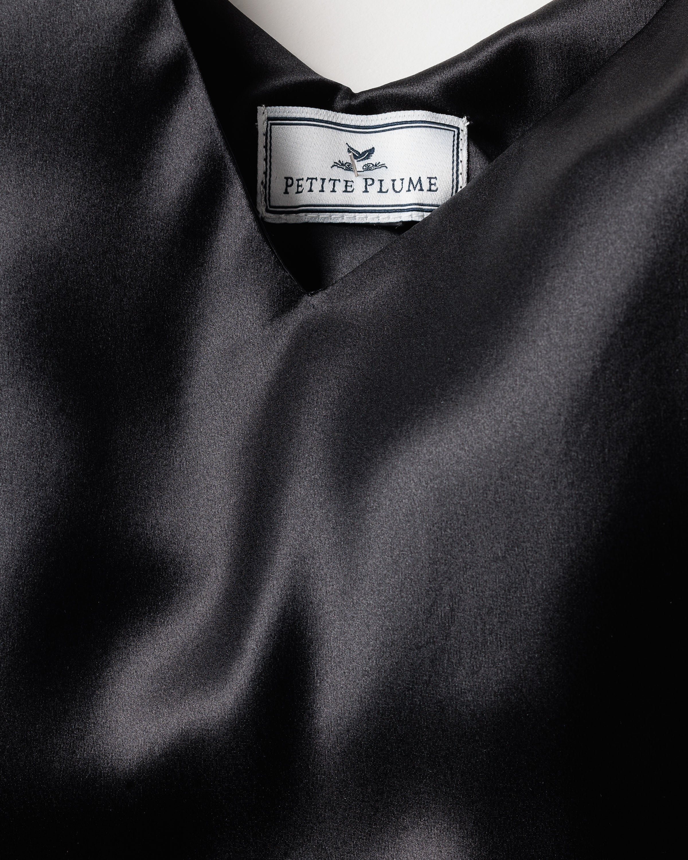 The smooth, shiny black fabric, resembling luxurious mulberry silk, features a visible label with Petite Plume in black font on white. It is softly folded and part of the Womens Silk Long Slip collection in Black by Petite Plume.