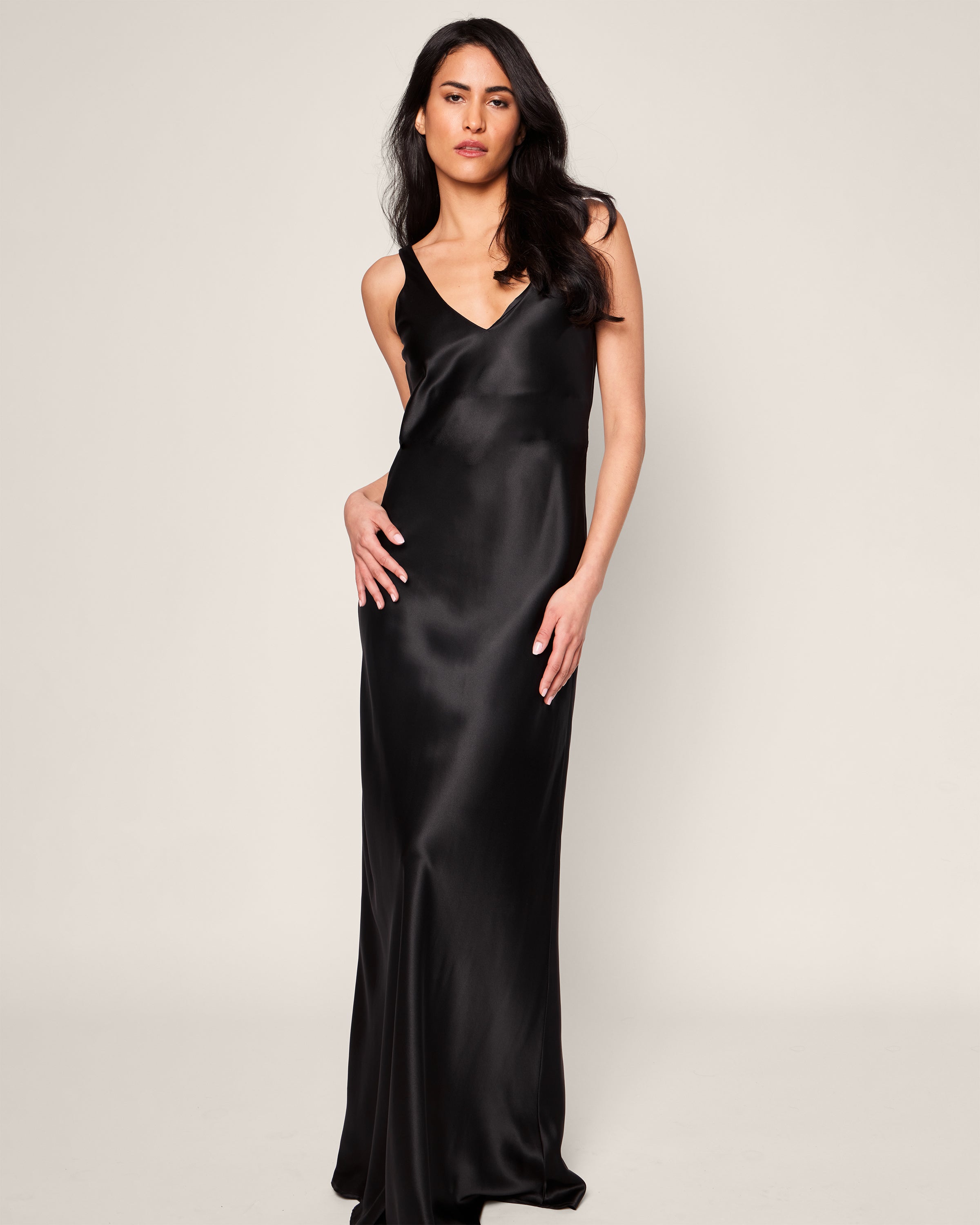 A woman poses against a plain backdrop, elegantly donning the Womens Silk Long Slip in Black by Petite Plume. The dress has a V-neckline and thin straps. Her black hair falls in gentle waves past her shoulders as she gazes at the camera with her right hand resting on her hip.