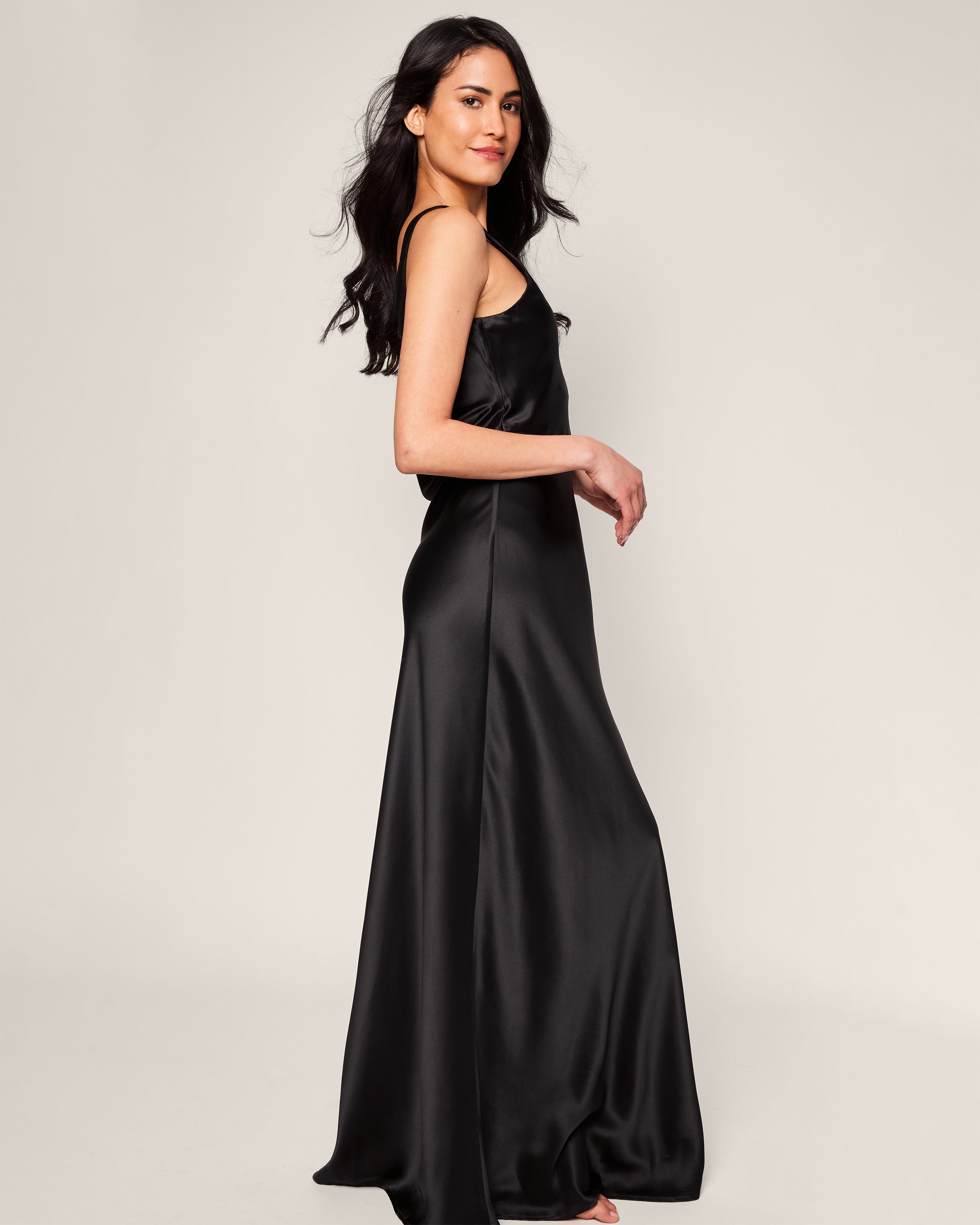 A person in a sleeveless Womens Silk Long Slip in Black by Petite Plume stands against a plain white background, smiling slightly with long hair cascading over their shoulder. Designed from exquisite mulberry silk, it glides effortlessly as they face sideways, looking towards the camera.
