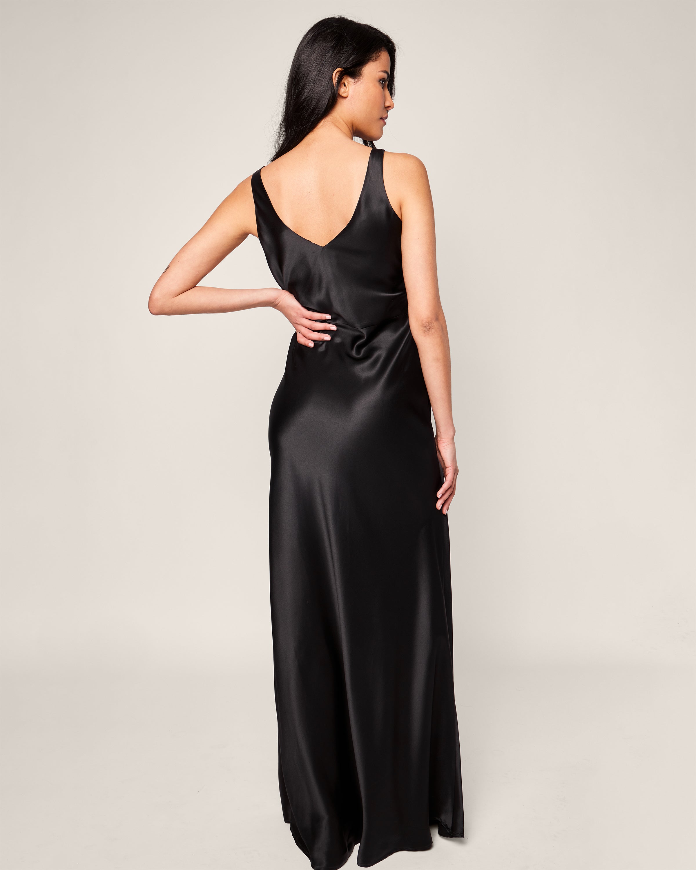 A woman in a Womens Silk Long Slip by Petite Plume, styled like luxurious sleepwear, poses with her back to the camera and hand on hip against a neutral background.