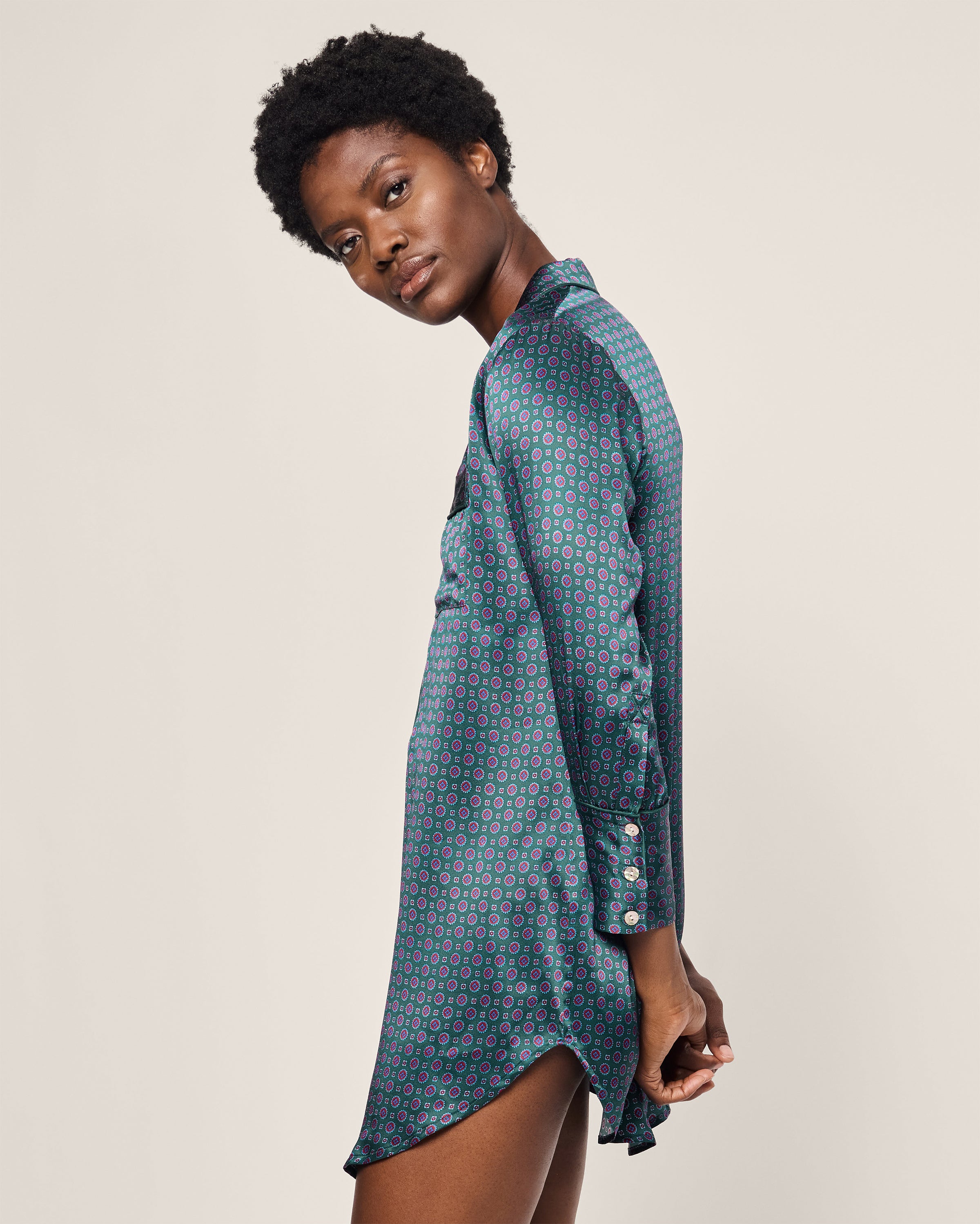 A woman with short curly hair is wearing a dark blue Womens Silk Nightshirt in Emerald Foulard from Petite Plume, featuring green and pink patterns. She stands sideways, slightly turning her head toward the camera against a plain, light-colored background.