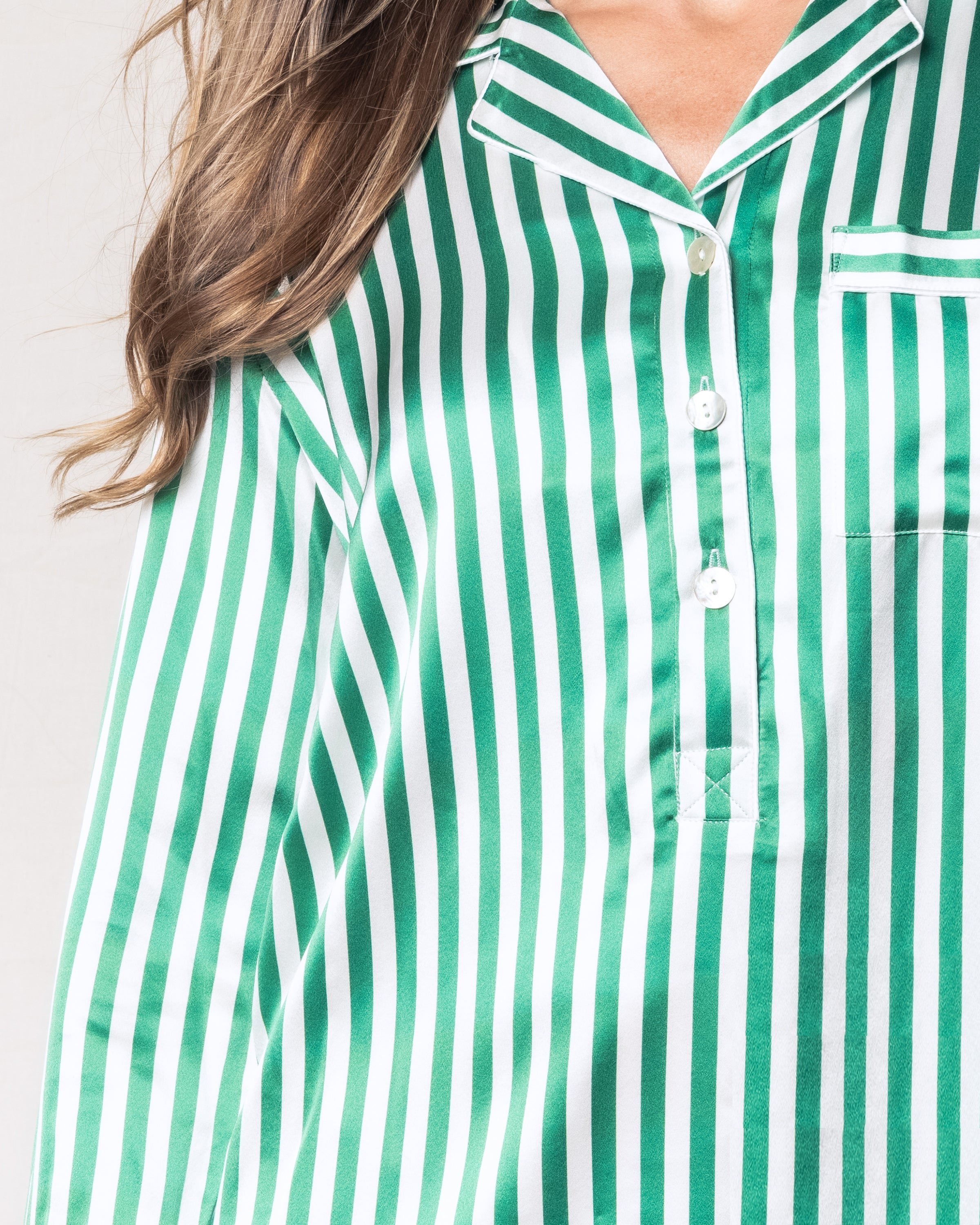 A close-up shows a person wearing Petite Plumes Womens Silk Nightshirt in Green Stripe, featuring an emerald green vertical stripe and a small left-side pocket. The Mulberry Silk fabric has a smooth, satin-like texture, with the persons hair cascading over the right shoulder.