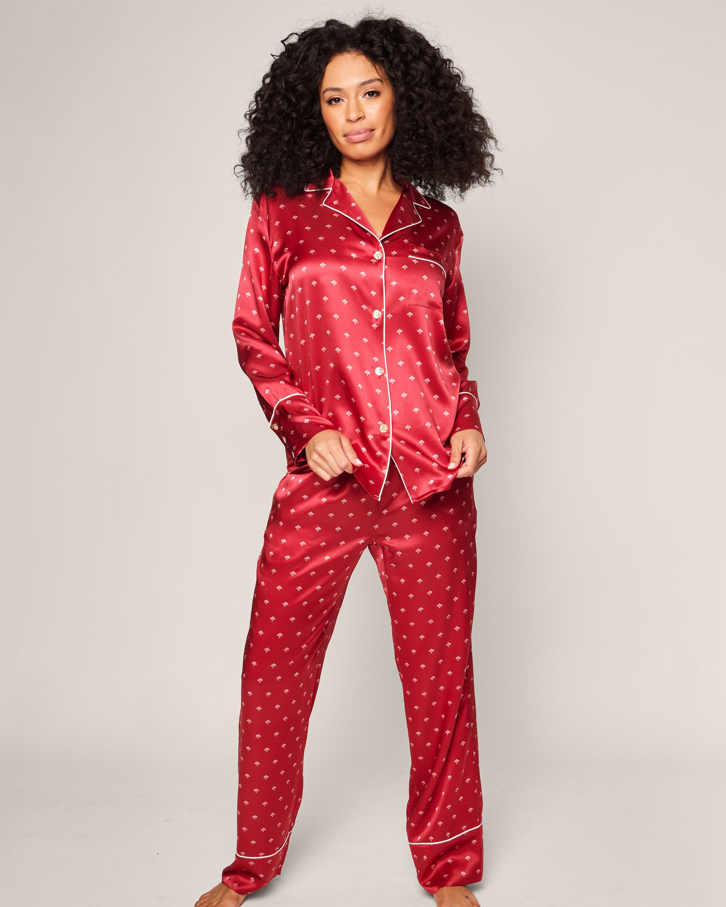 A person in a Crimson Art Nouveau Womens Silk Pajama Set by Petite Plume stands barefoot against a neutral backdrop. The set, made of luxurious mulberry silk, includes a button-up top with a collar and matching pants. Their curly hair frames their relaxed, serene expression.