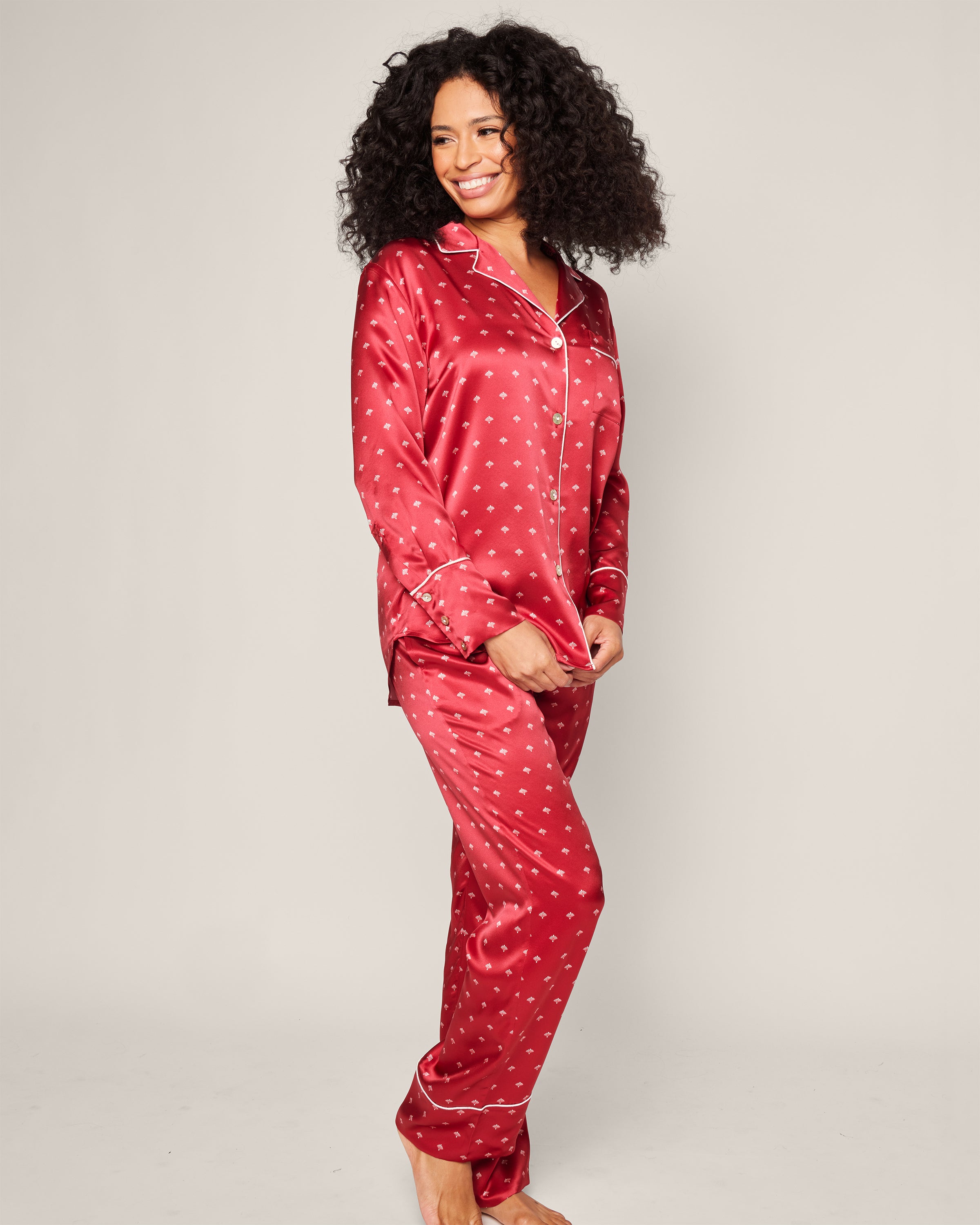 A woman with curly hair smiles warmly, wearing Petite Plumes Womens Silk Pajama Set in Crimson Art Nouveau. Her ensemble exudes the elegance of luxury sleepwear as she stands gracefully against a plain background.