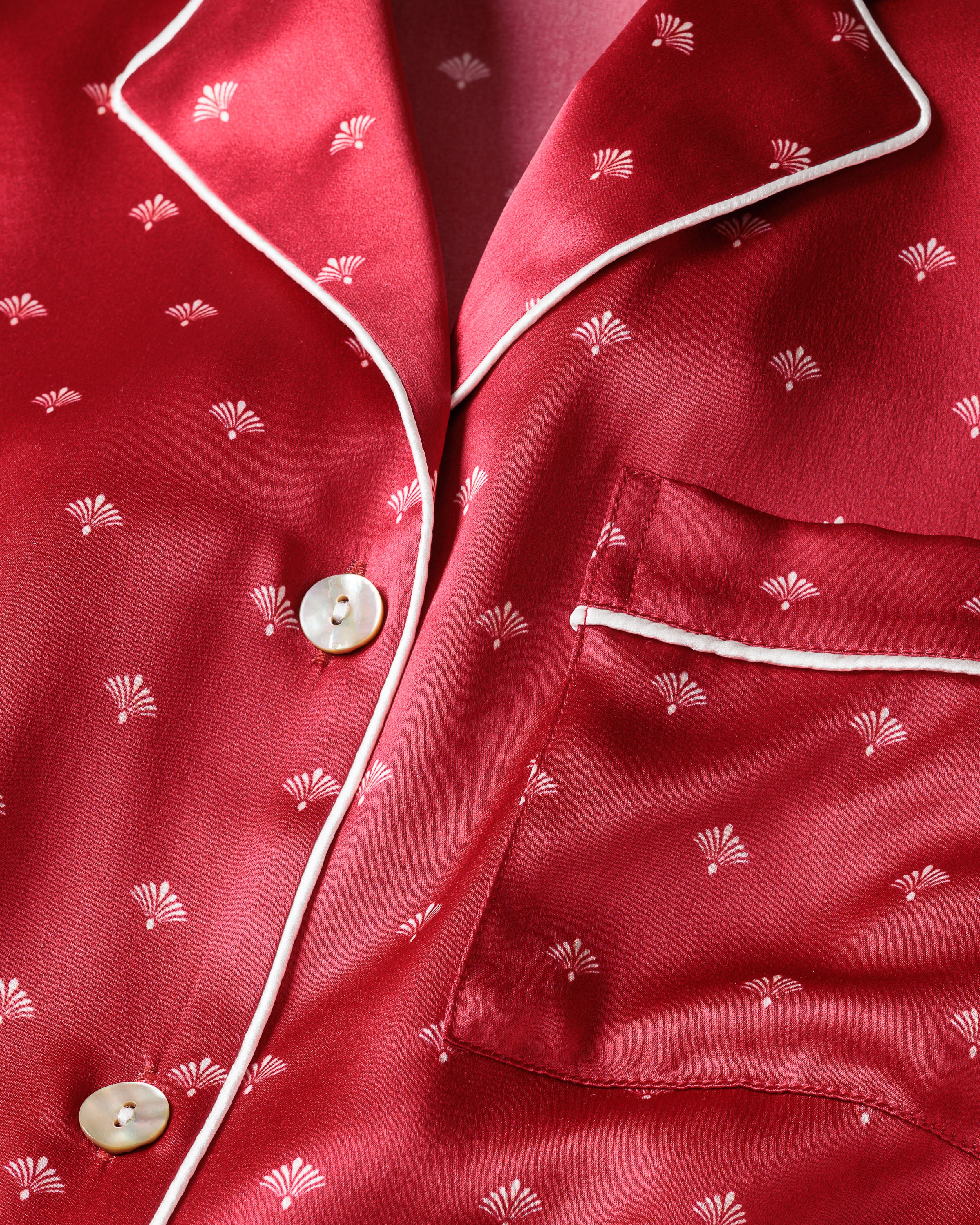 Close-up of the Womens Silk Pajama Set in Crimson Art Nouveau by Petite Plume, featuring a luxurious red mulberry silk top with subtle white floral patterns. Elegantly finished with white piping on the collar and pocket, it is fastened with delicate white buttons.
