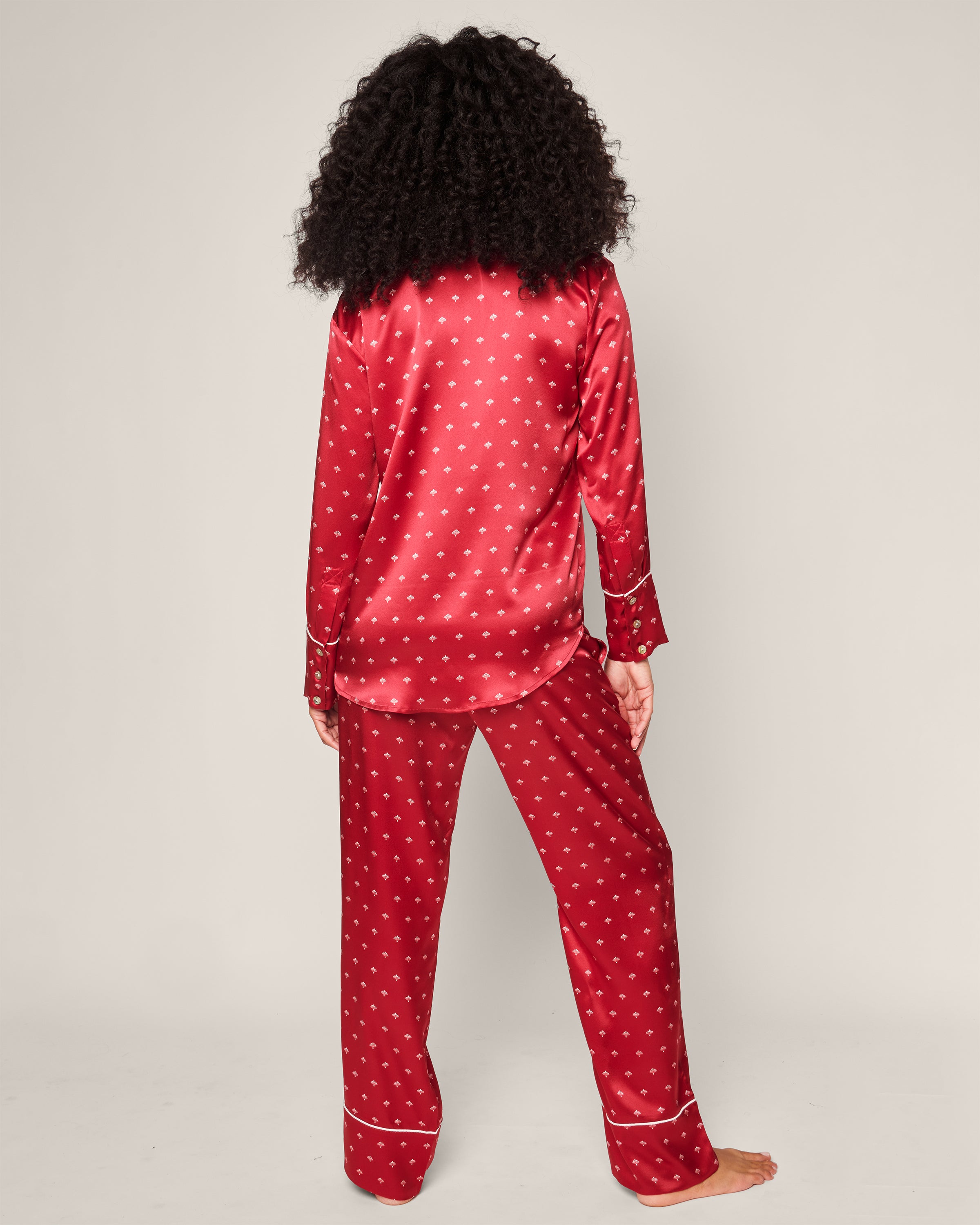 A person with curly hair stands barefoot facing away, wearing Petite Plumes Womens Silk Pajama Set in Crimson Art Nouveau. The mulberry silk set is adorned with small polka dots and elegant white piping. A plain background highlights the opulence of this luxurious sleepwear.