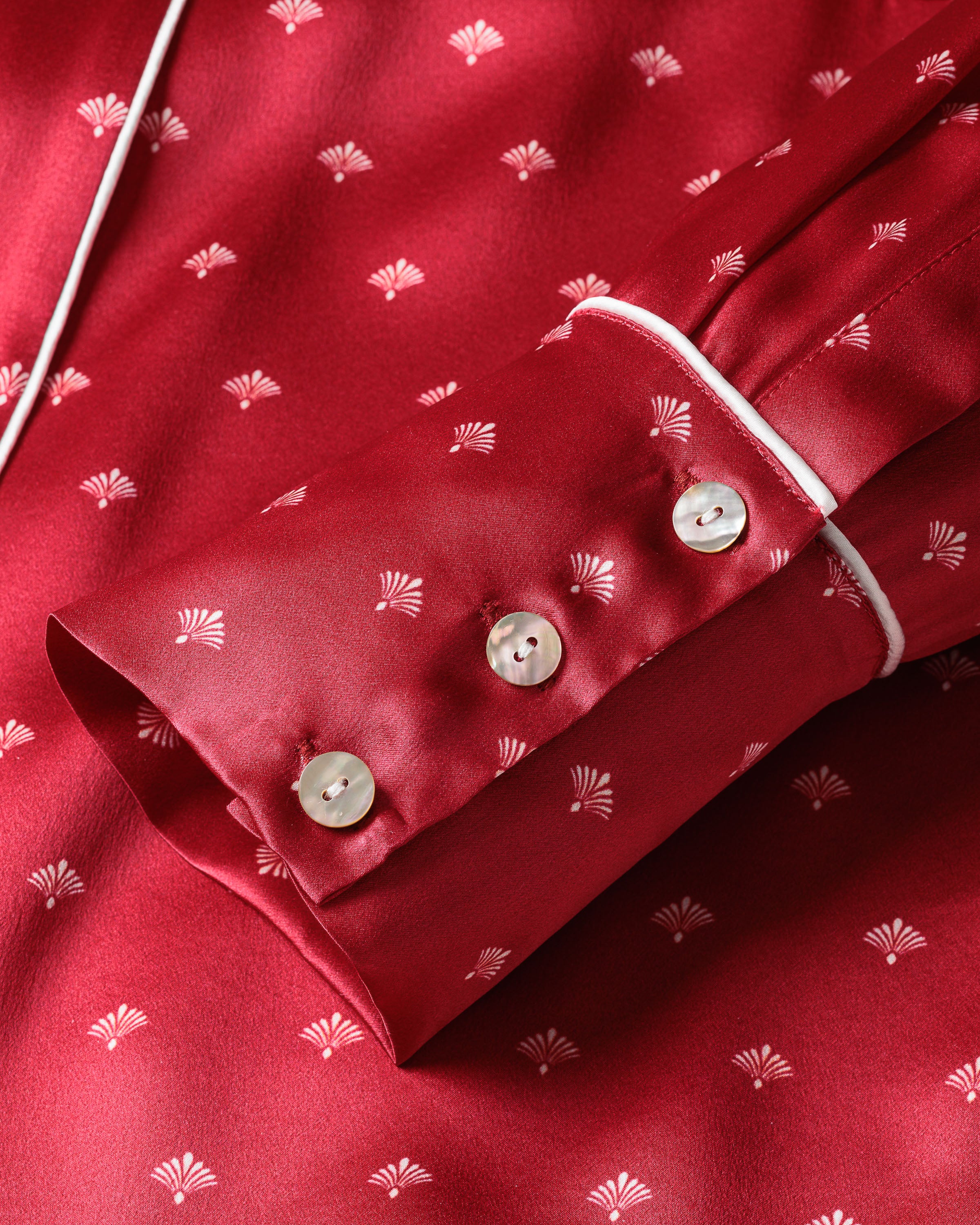 A close-up of Petite Plumes Womens Silk Pajama Set in Crimson Art Nouveau showcases the fabrics small white fan patterns. The sleeve cuff, adorned with white piping and three buttons, embodies the elegance of luxury sleepwear.