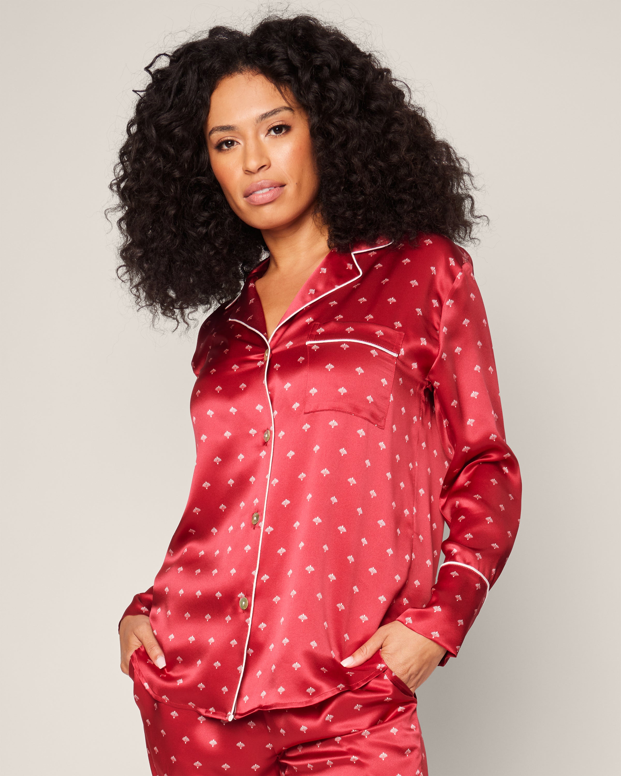 A person with curly hair wears the Womens Silk Pajama Set in Crimson Art Nouveau from Petite Plume, akin to luxury sleepwear with a subtle white pattern. The shirt has a pocket and white piping. They pose with one hand in their pocket against a light gray background.