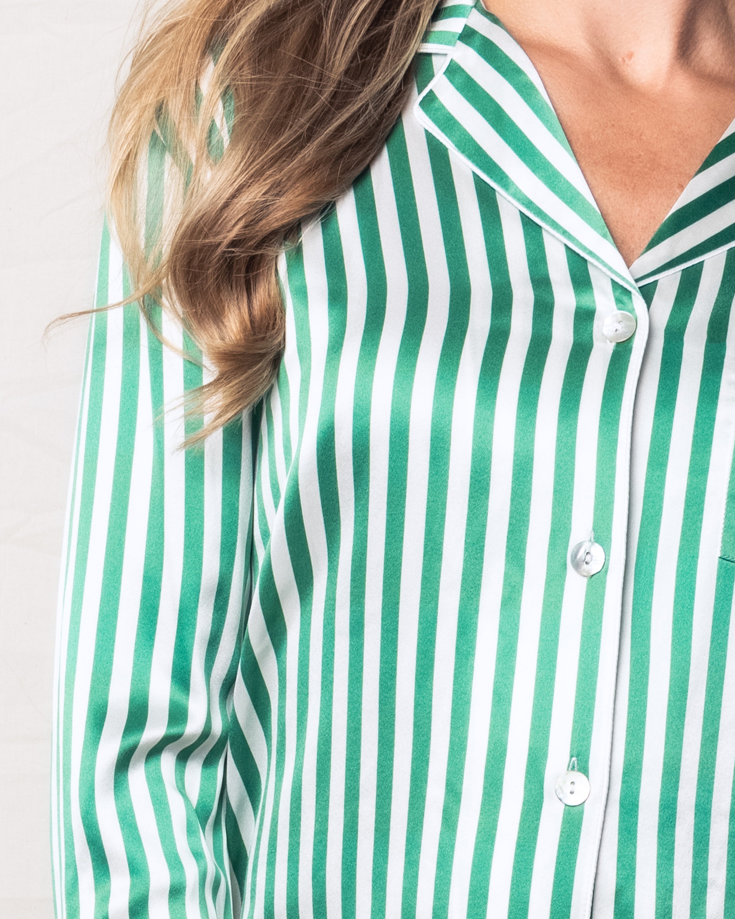 The image depicts a person in Petite Plumes Womens Silk Pajama Set in Green Stripe, featuring a collar, button-up front, and visible pocket. Their long hair elegantly drapes over the left shoulder.