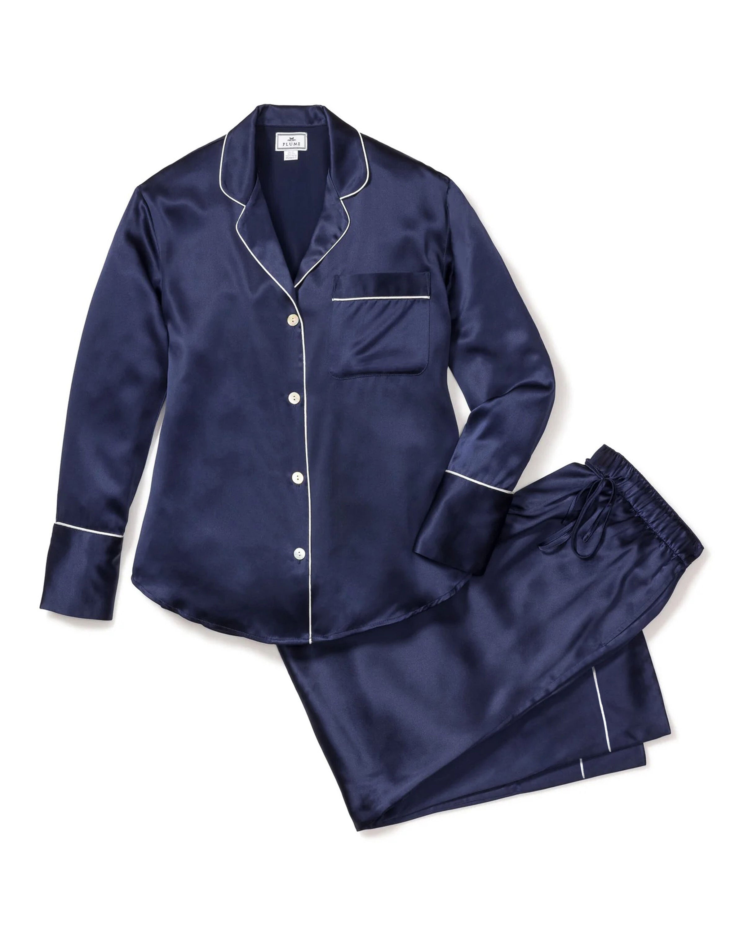A person stands in Petite Plumes Womens Silk Pajama Set in Navy, showcasing luxurious sleepwear with white piping and a classic design—featuring a long-sleeve button-up top with a pocket and matching pants—against a plain background.