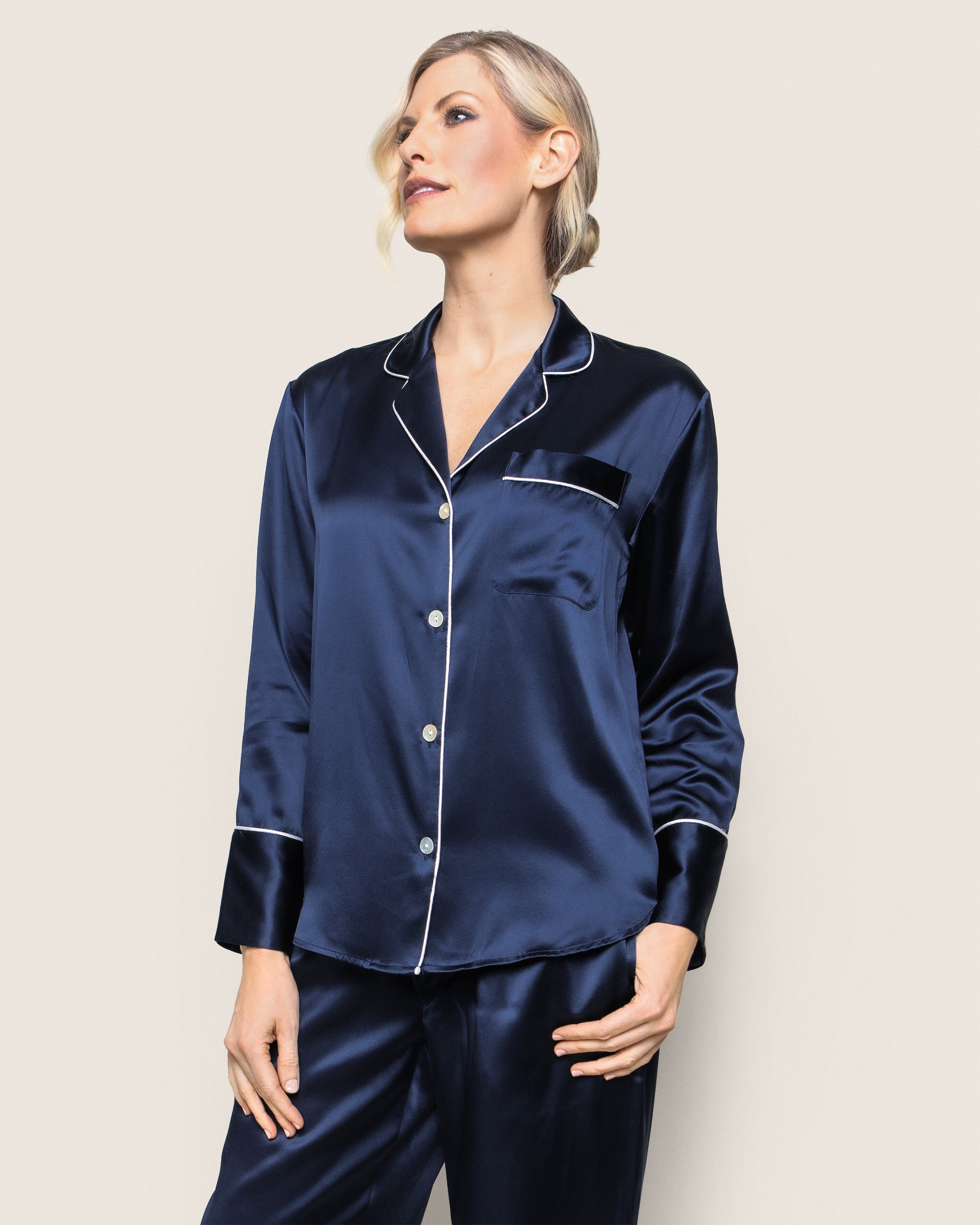 A person with short blonde hair stands against a light background wearing Petite Plumes Womens Silk Pajama Set in Navy. This luxury sleepwear has a timeless silhouette, white piping, and a breast pocket. They appear to be looking to the side.
