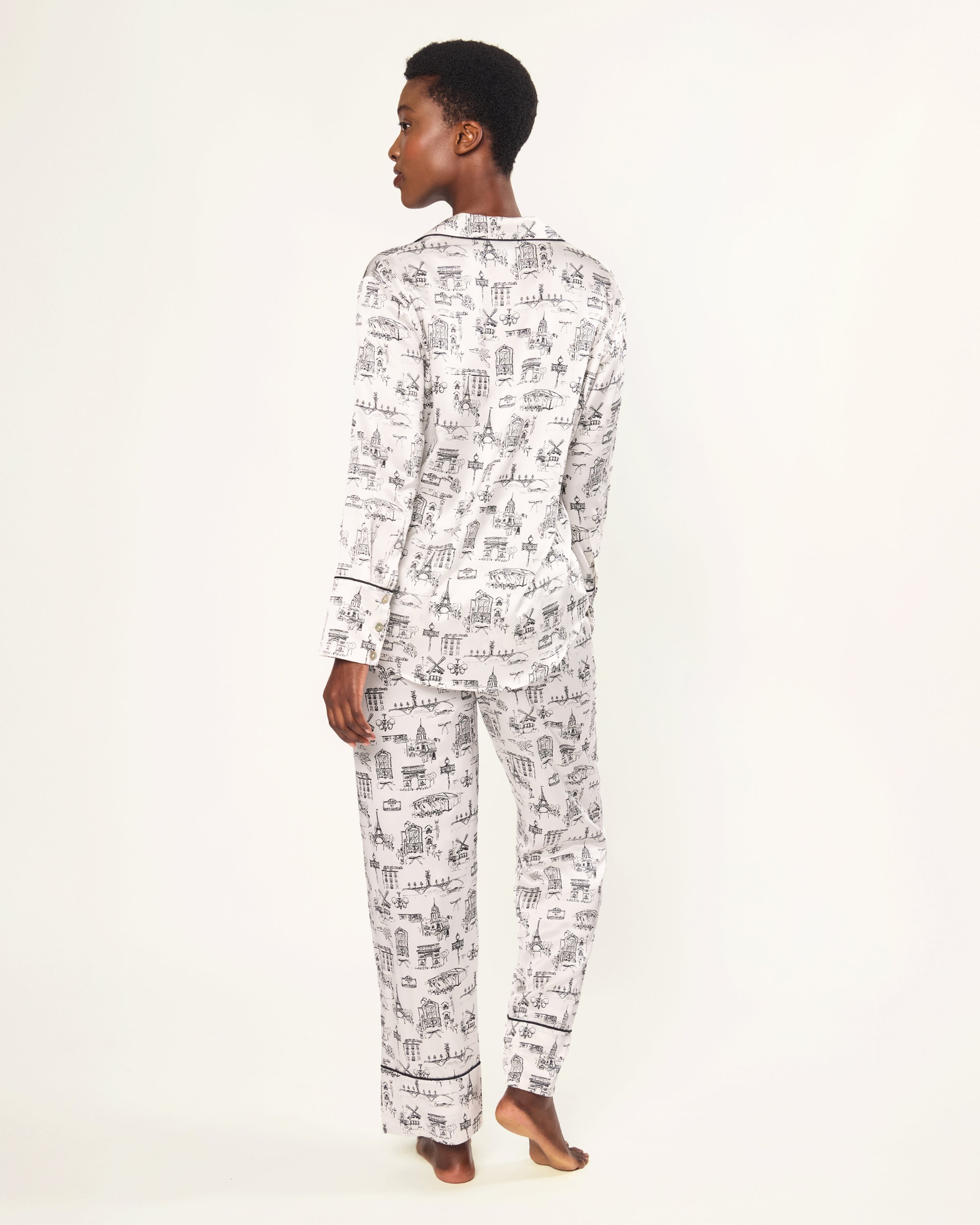 Women's Silk Pajama Set in Parisian Toile: white button-up with black piping, illustrated with buildings and trees. Includes long-sleeve top with chest pocket and matching long pants featuring an elastic waistband.
