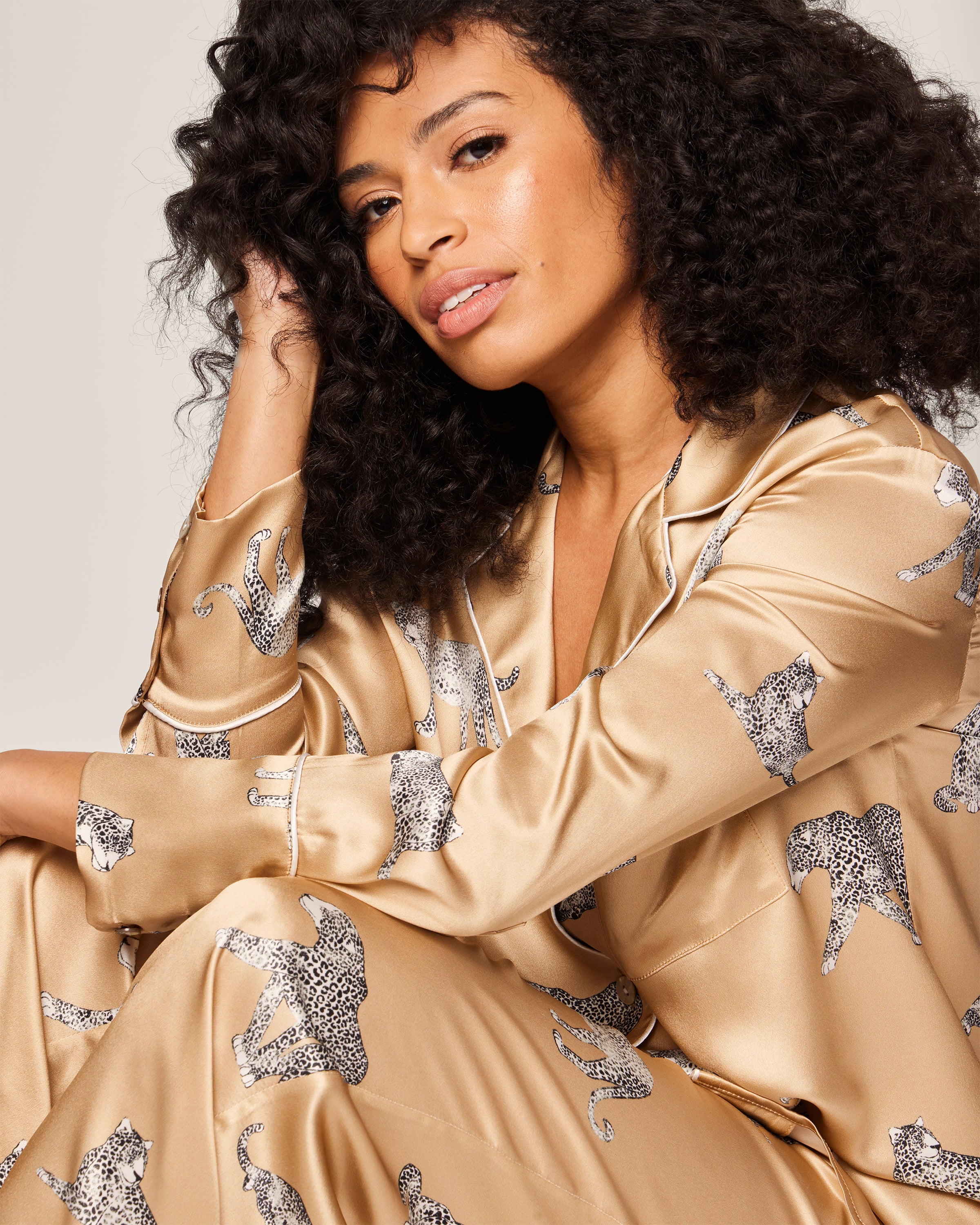 A woman with curly hair poses confidently in a Petite Plume Womens Silk Pajama Set in Panthère Champagne, featuring black and white panther motifs. Resting her head on her hand, she stands out against the plain backdrop, highlighting the luxurious sleepwear.
