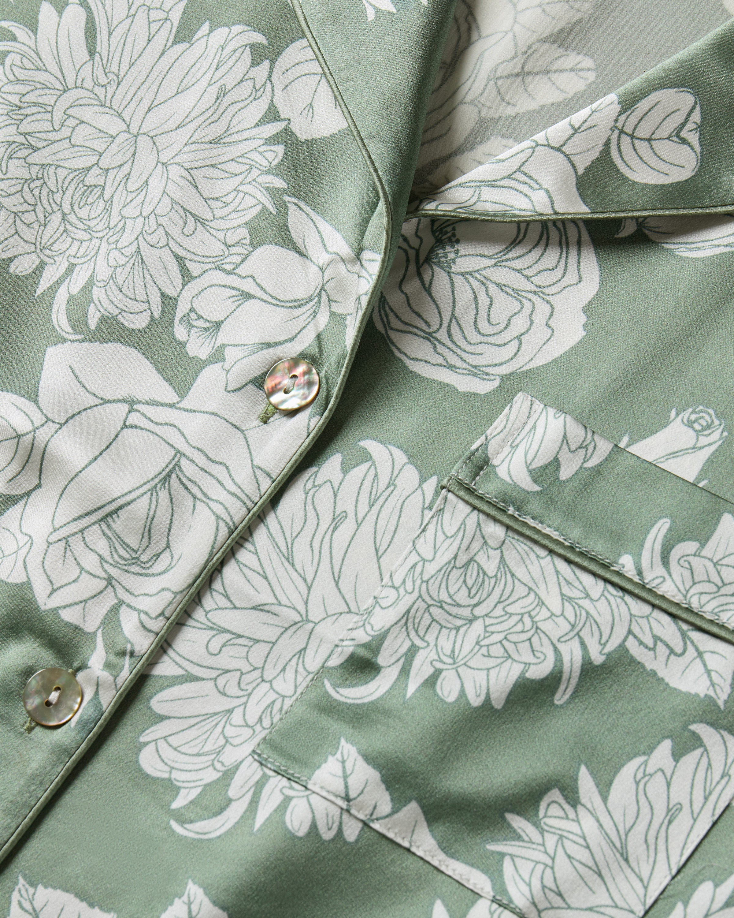 Women's Silk Pajama Set in Sage Bloom: a sage green satin set with a white floral pattern, featuring a long-sleeve button-up top with chest pocket and matching drawstring pants.