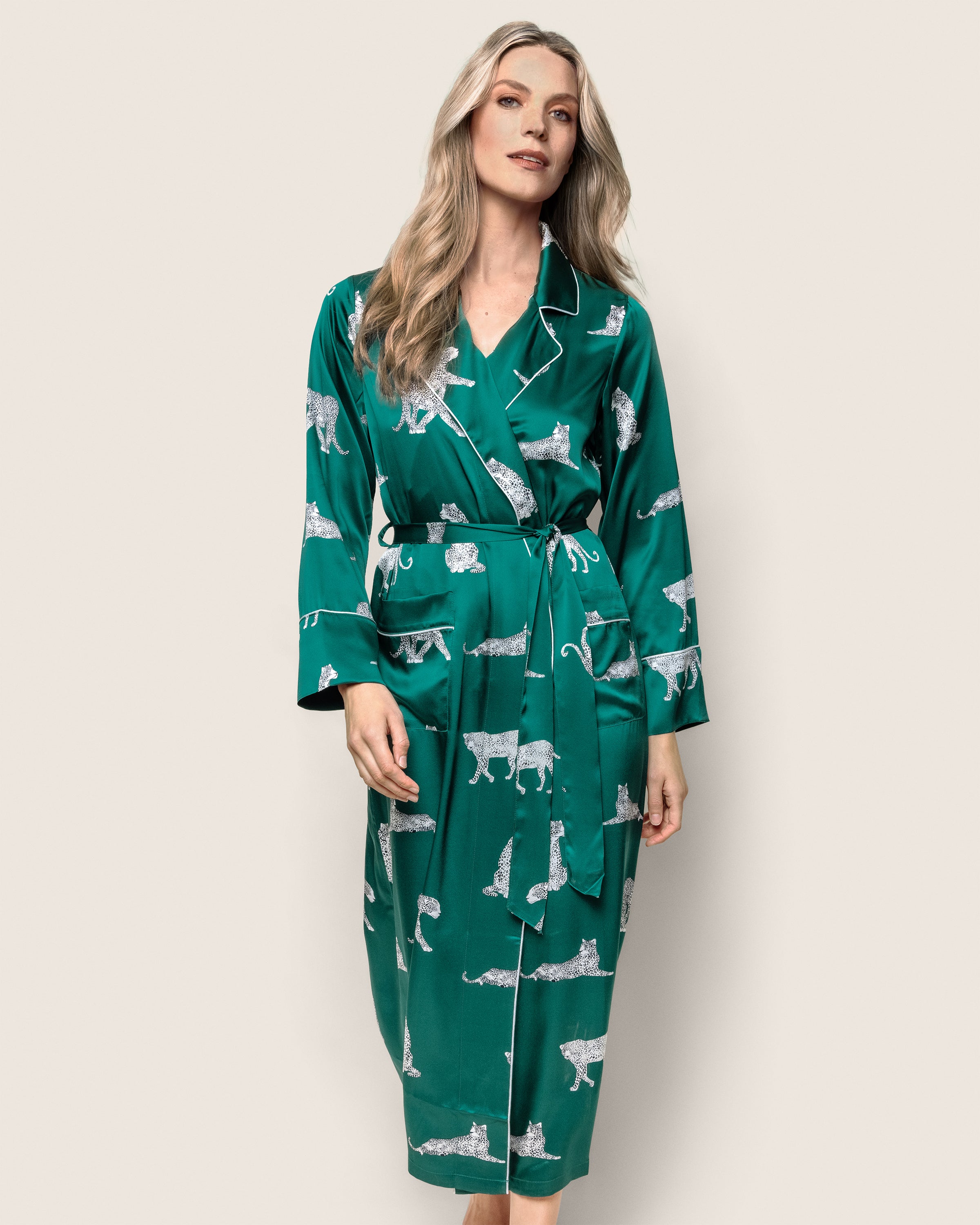 A person wears Petite Plumes Womens Silk Long Robe in Panthère de Luxe, a luxurious green robe featuring white leopard outlines. Adorned with piped edges, it includes a belt and long sleeves against a plain, light background.