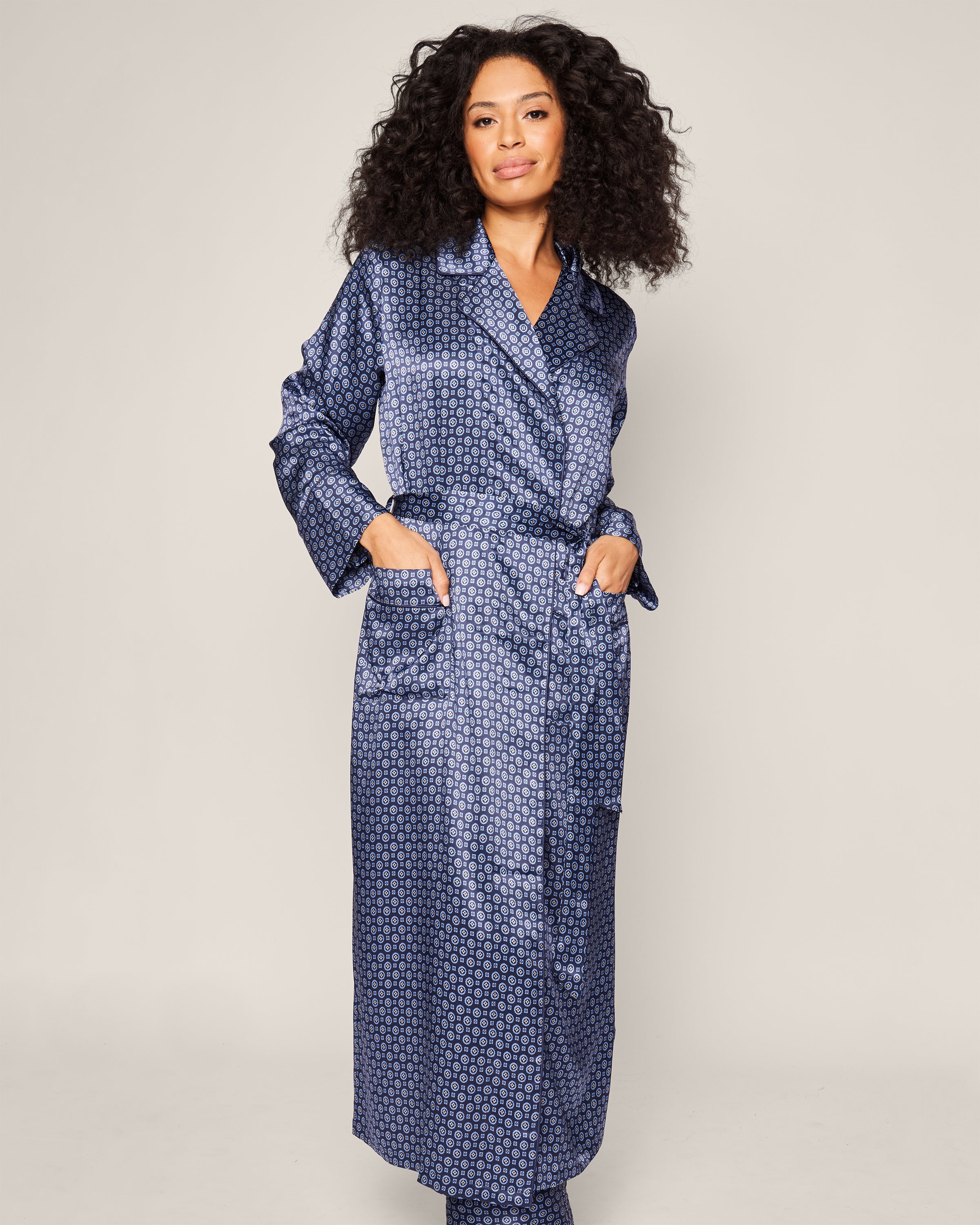 A person with curly hair smiles, wearing Petite Plume’s Womens Silk Robe in Midnight Foulard. This mulberry silk robe, adorned with dots, includes a waist tie and pockets. They stand against a plain light background, epitomizing comfort and style.