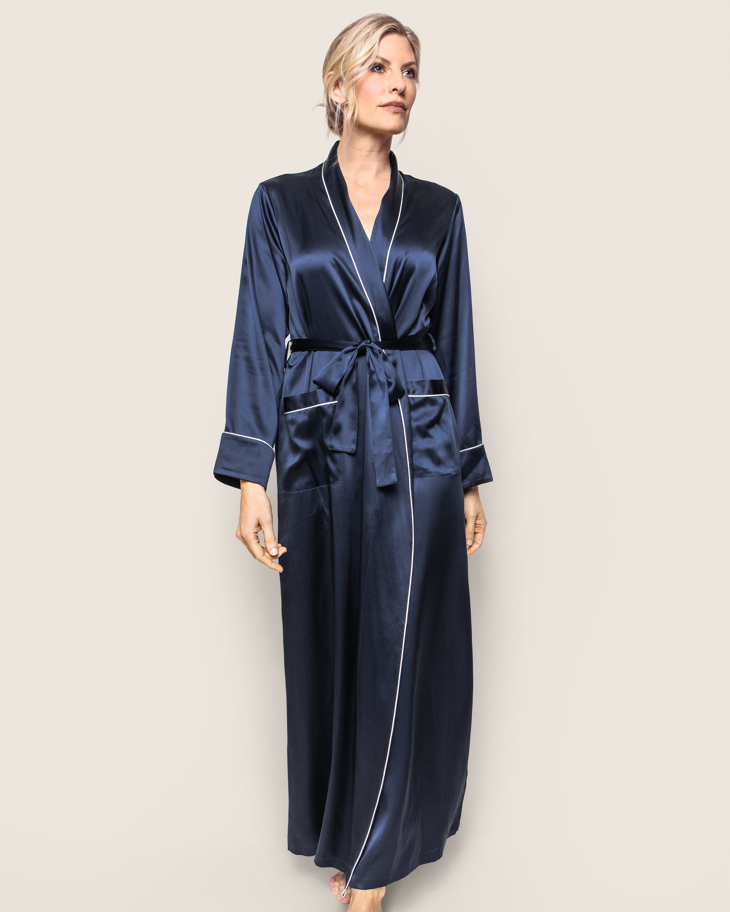 Dressed in a Petite Plume Womens Silk Long Robe in Navy, featuring white piping and a tied waist belt, a person stands against a light background. Made from 22mm silk, their blonde hair is styled up as they look slightly to the side.
