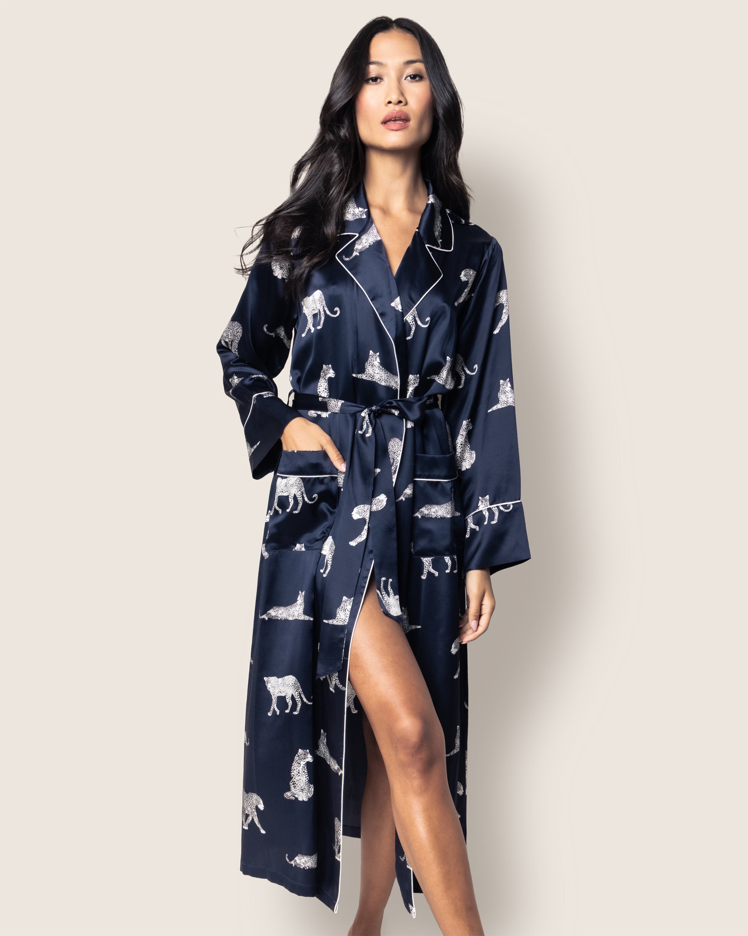 A woman poses confidently in Petite Plumes Womens Silk Long Robe in Panthère de Nuit, a luxury animal print satin robe with a belt tie. She stands against a beige background, exuding classic Hollywood glamour, with one hand stylishly tucked in her pocket.