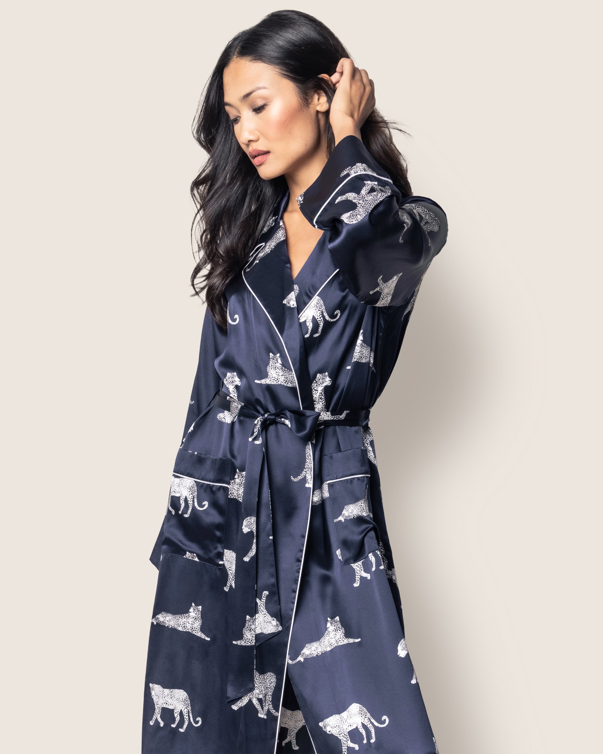 A person with long dark hair wears Petite Plumes Womens Silk Long Robe in Panthère de Nuit. The navy robe, adorned with white animal prints and tigers, features a waist tie and piped edges. They pose against a plain light background, gently touching their hair in classic Hollywood style.