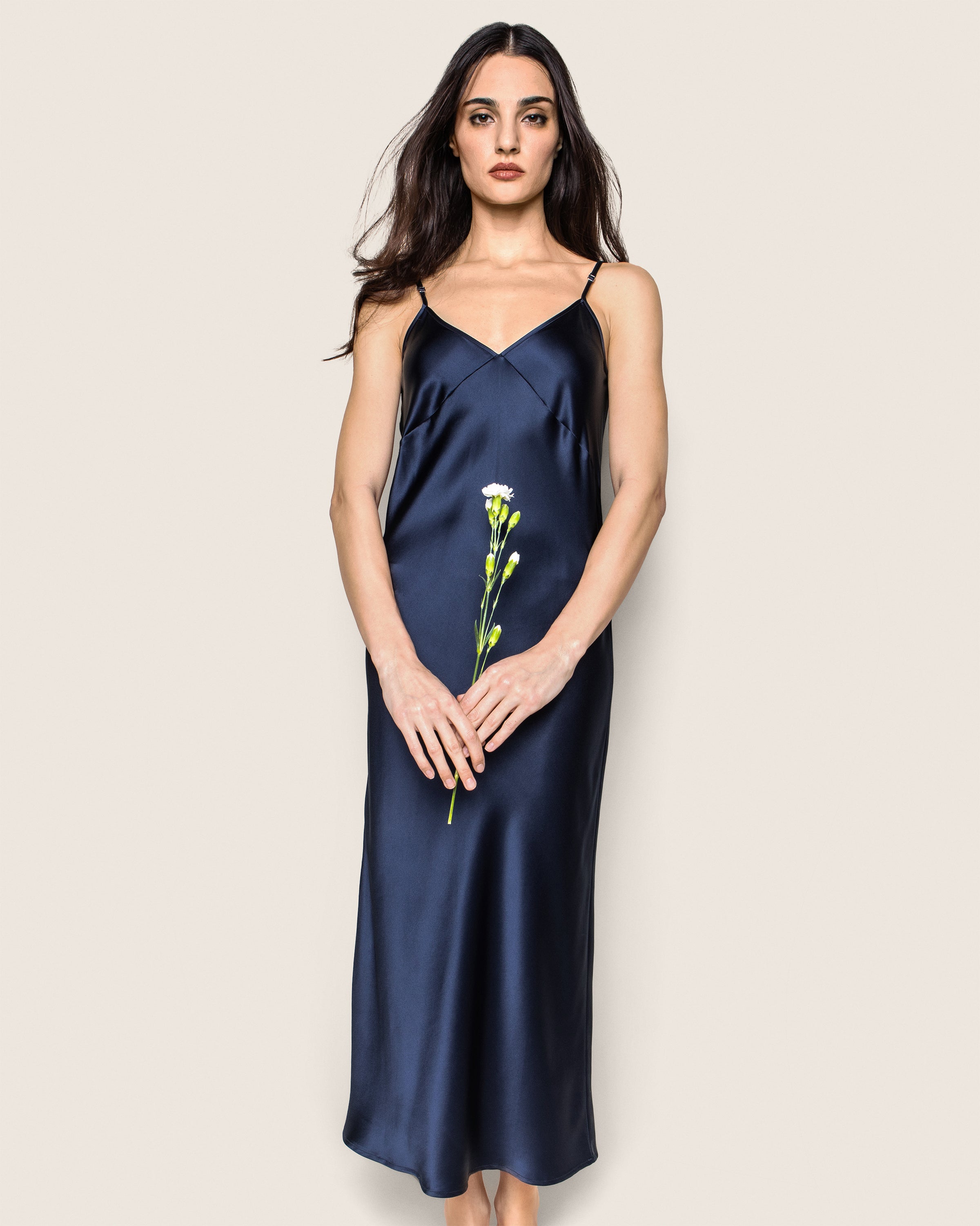 A woman in a Petite Plume Womens Silk Cosette Nightgown in Navy stands against a neutral backdrop, holding a single white flower with both hands. Her hair is down as she gazes forward calmly.