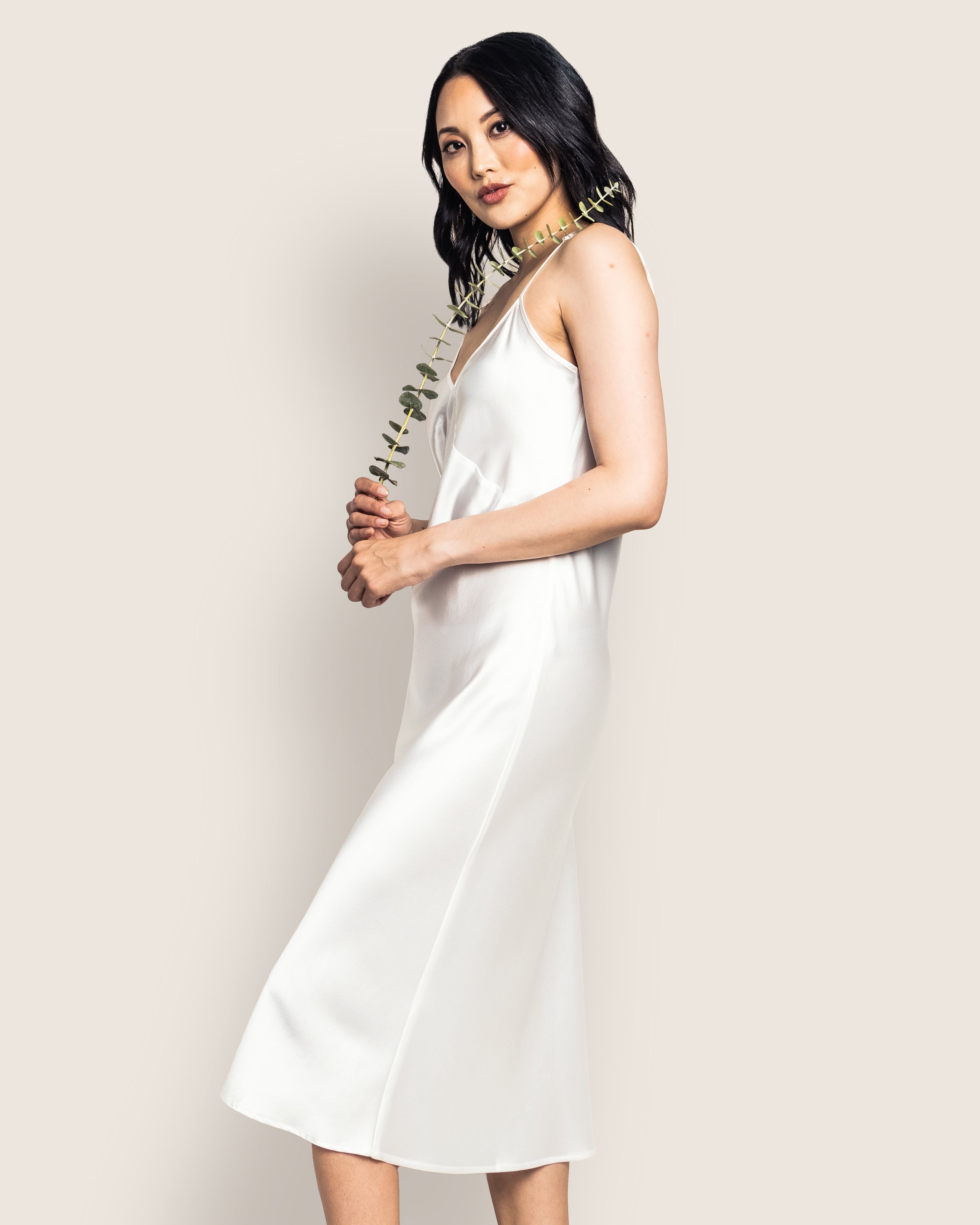 A woman in the Petite Plume Womens Silk Cosette Nightgown in White stands sideways against a plain background, holding a branch with green leaves. With loose dark waves framing her face, she gazes at the camera, exuding elegance and serenity.