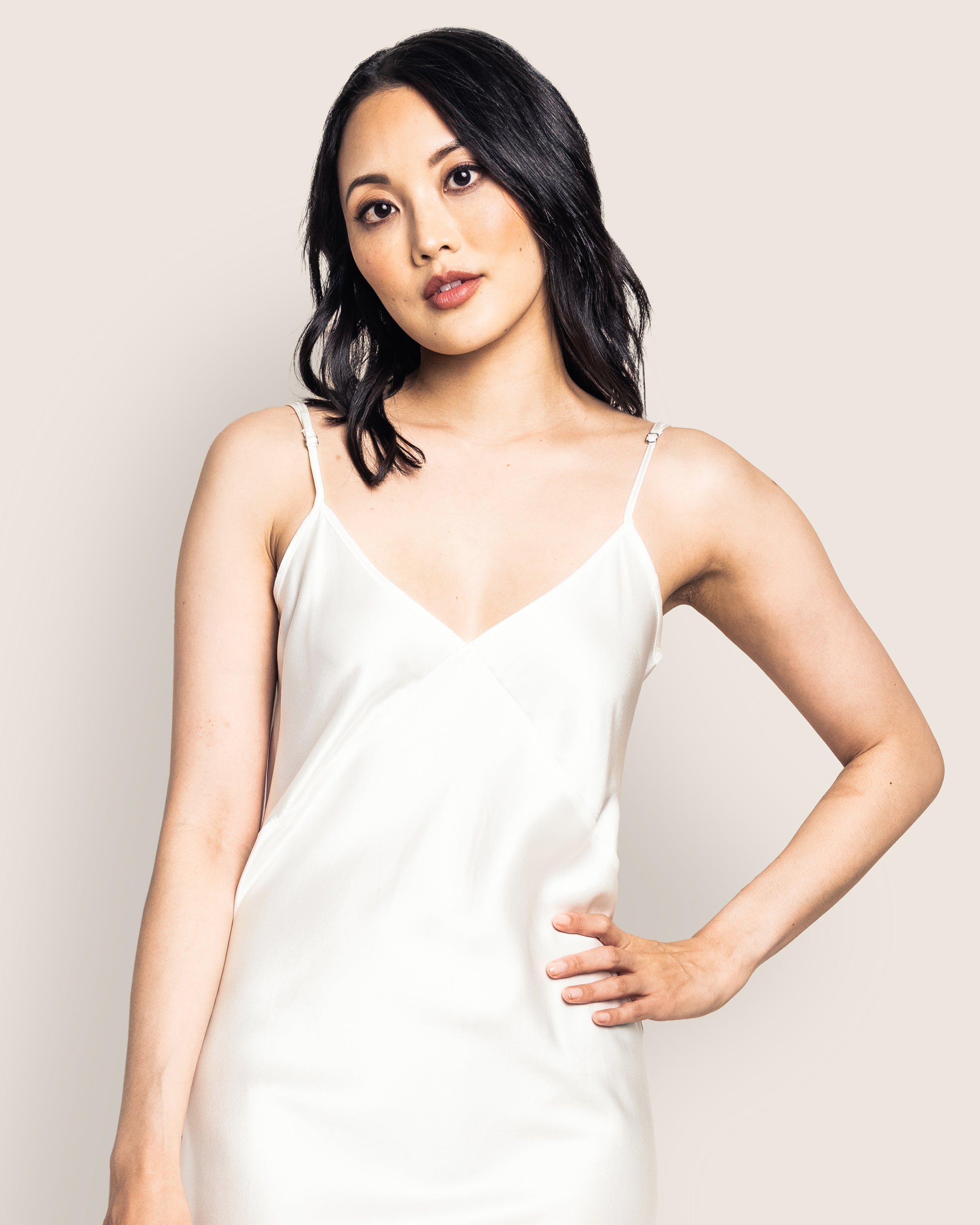 A woman with long, wavy black hair poses elegantly in the Womens Silk Cosette Nightgown by Petite Plume. She stands with one hand on her hip against a light beige background, looking directly at the camera with a calm expression.