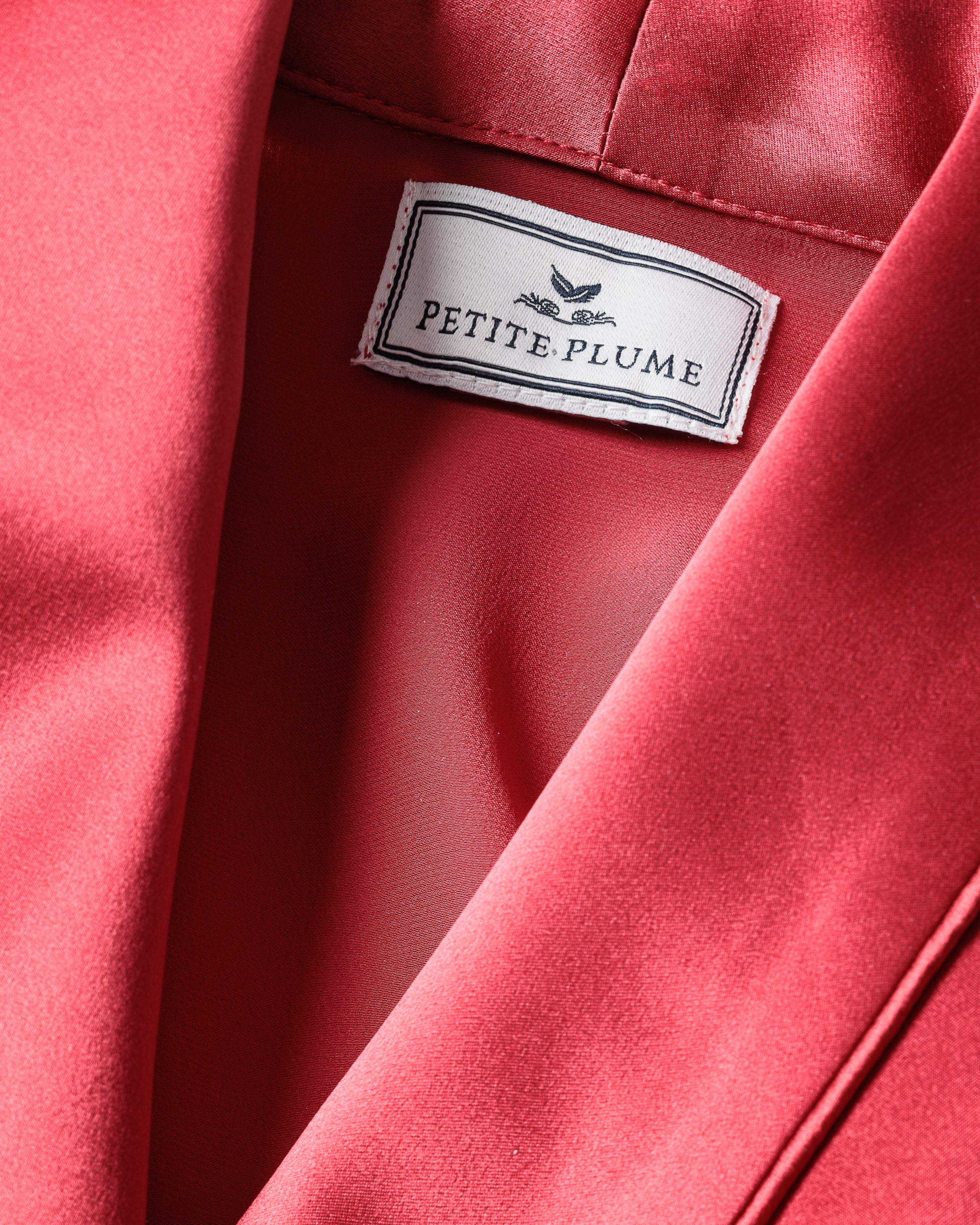 Close-up of a crimson satin fabric resembling luxurious mulberry silk with Petite Plume label sewn inside. Its smooth, shiny texture enhances its allure, ideal for elegant sleepwear thats stylish and hypoallergenic.