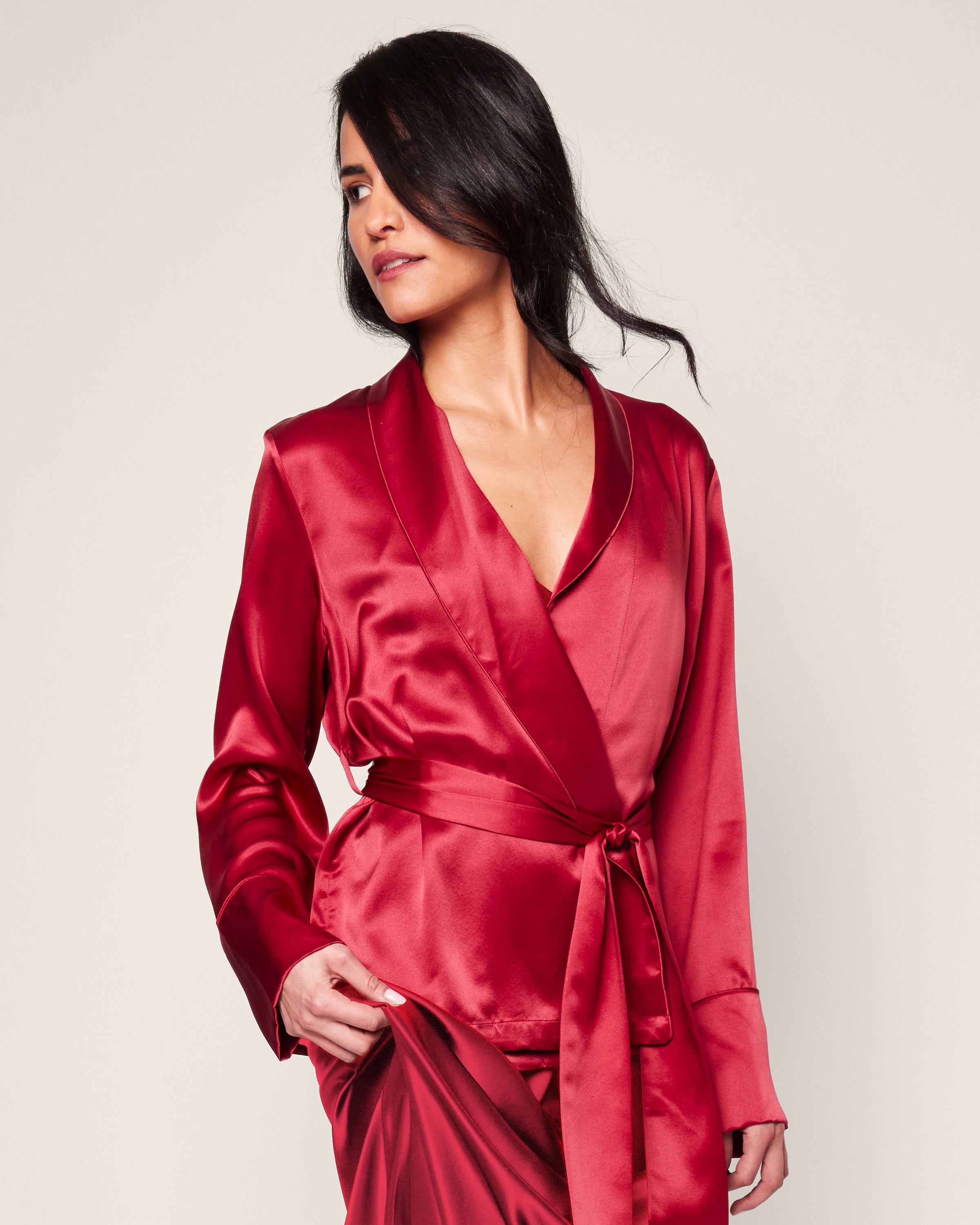 A woman models the Womens Silk Short Robe in Crimson by Petite Plume, with a matching sash. Her long dark hair is styled to one side against a plain backdrop, highlighting her chic attire reminiscent of elegant sleepwear crafted from hypoallergenic mulberry silk.
