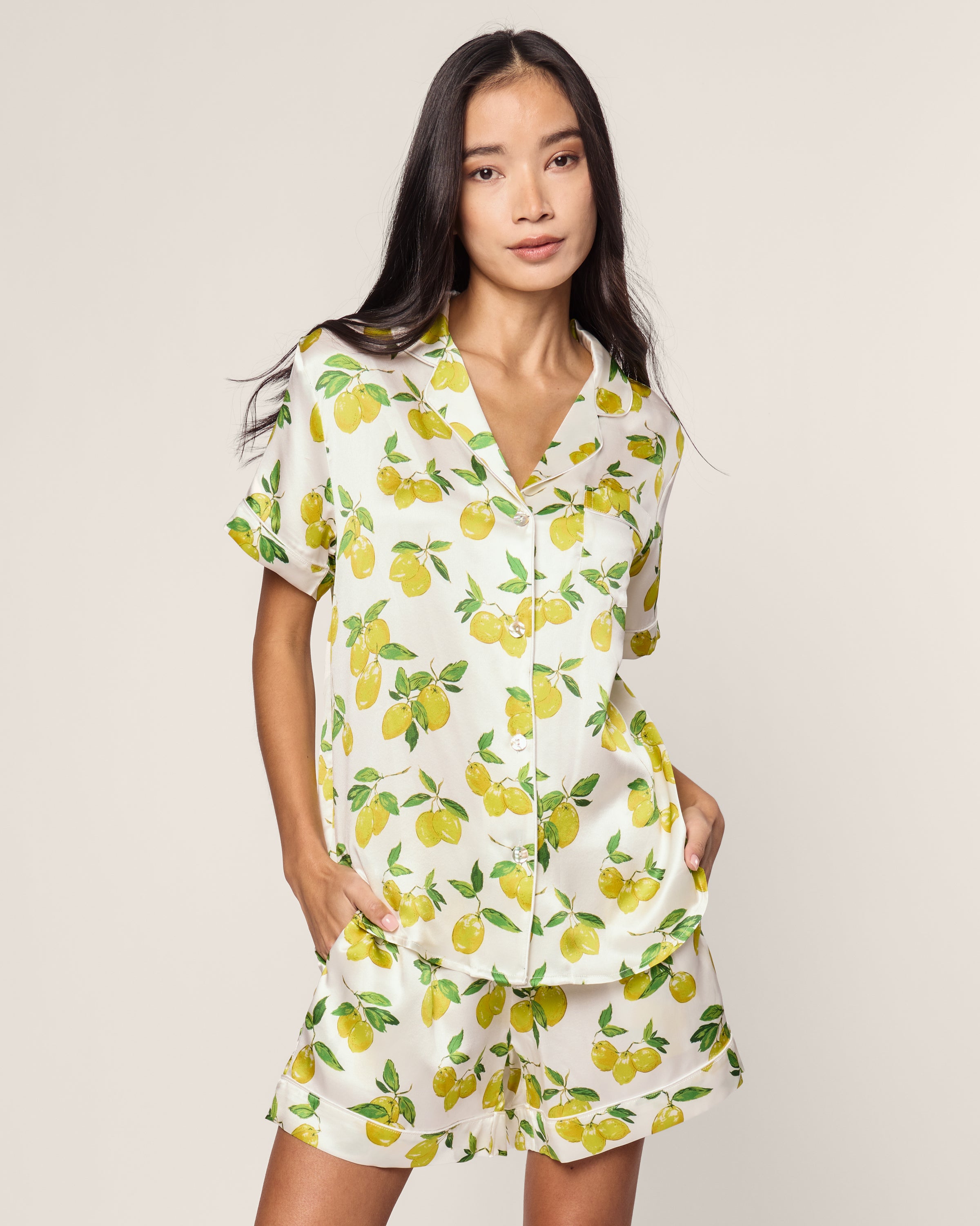The Womens Silk Classic Short Set in Amalfi Lemons by Petite Plume epitomizes elegance with its limited-edition design. The white short-sleeve button-up shirt and matching shorts feature vibrant yellow and green lemons, radiating effortless relaxation as she stands hands in pockets against a plain backdrop.