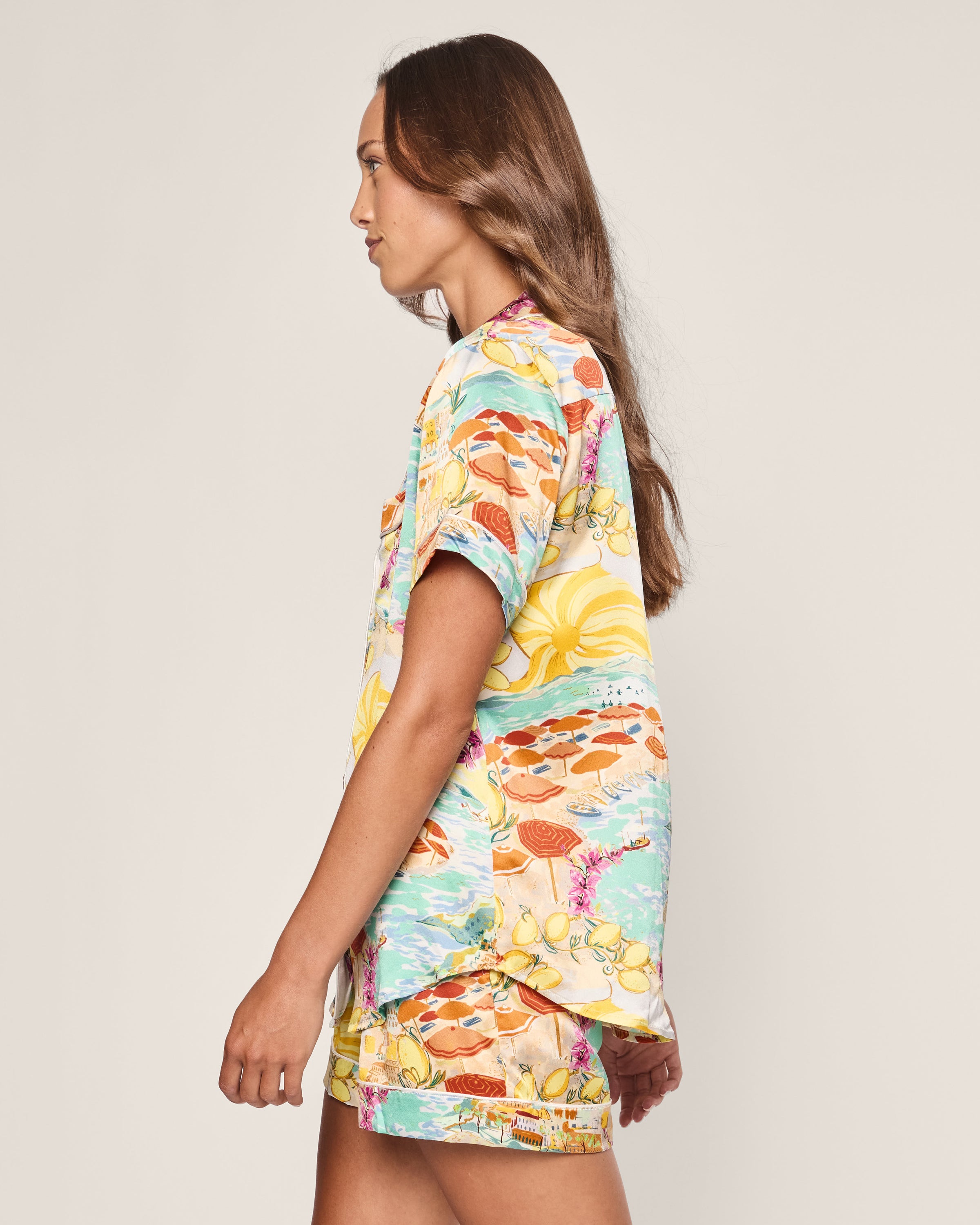 A woman in the Petite Plume Womens Silk Short Set in La Dolce Vita stands against a plain background. Facing left, her long hair flows down her back, while the vibrant design with abstract shapes and hints of lemon print evokes Italian seaside luxury.