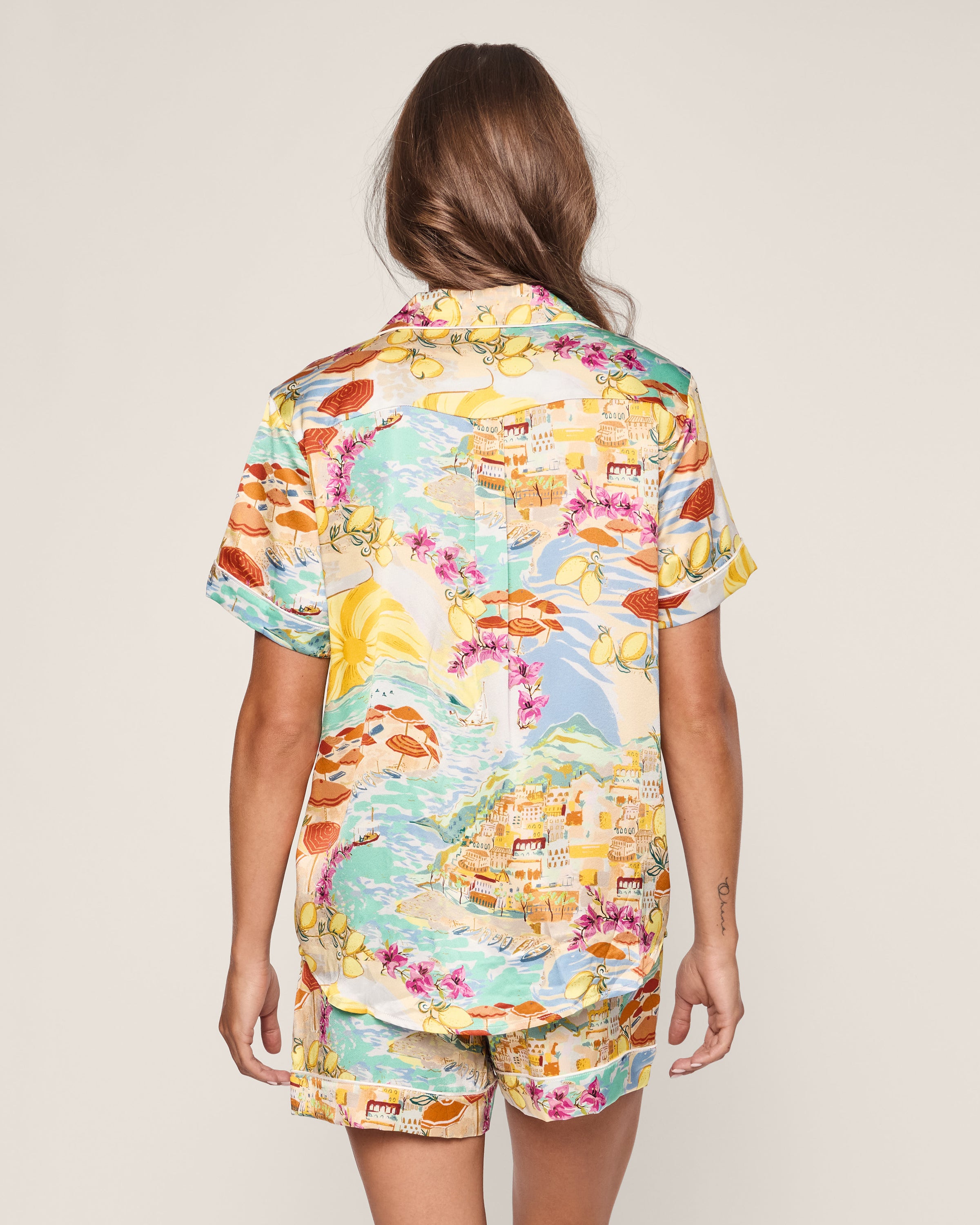 A person with long hair faces away, wearing Petite Plumes Womens Silk Short Set in La Dolce Vita. The colorful coastal-themed design of boats, flowers, and buildings evokes an Italian seaside feel against a plain backdrop, highlighting its vibrant pattern and leisurely luxury.